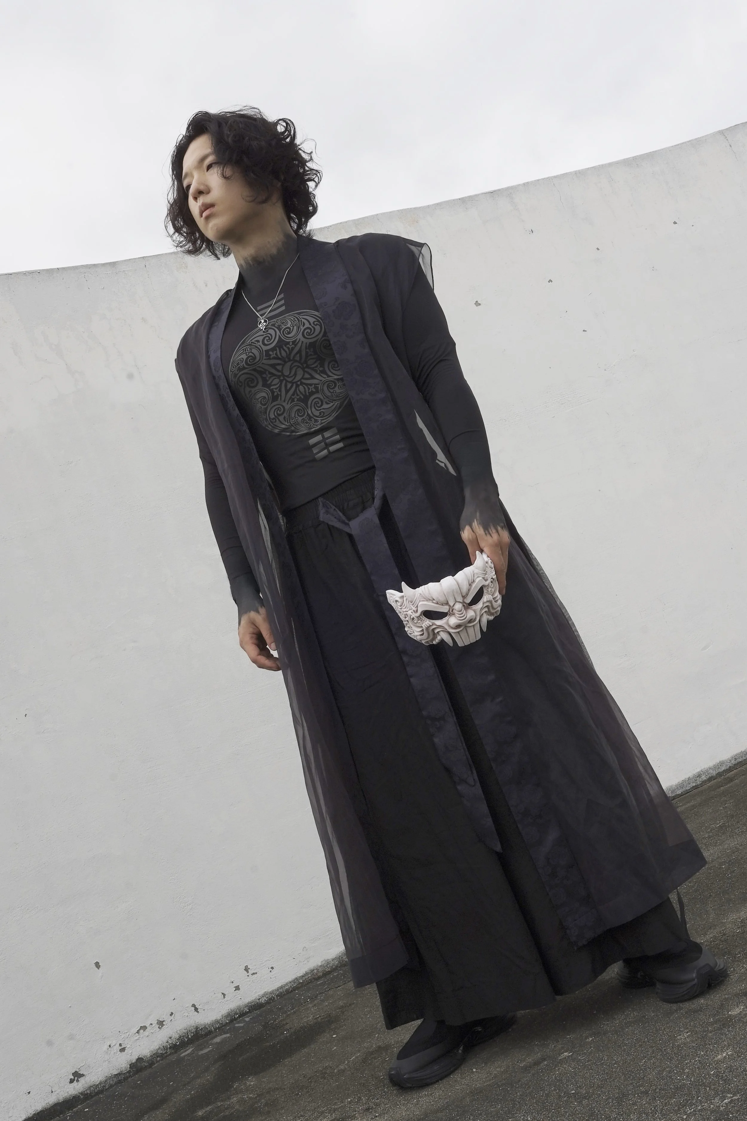 A person with long curly hair wearing black clothing and a black sheer cloak, holding a white decorative mask in front of a white wall.