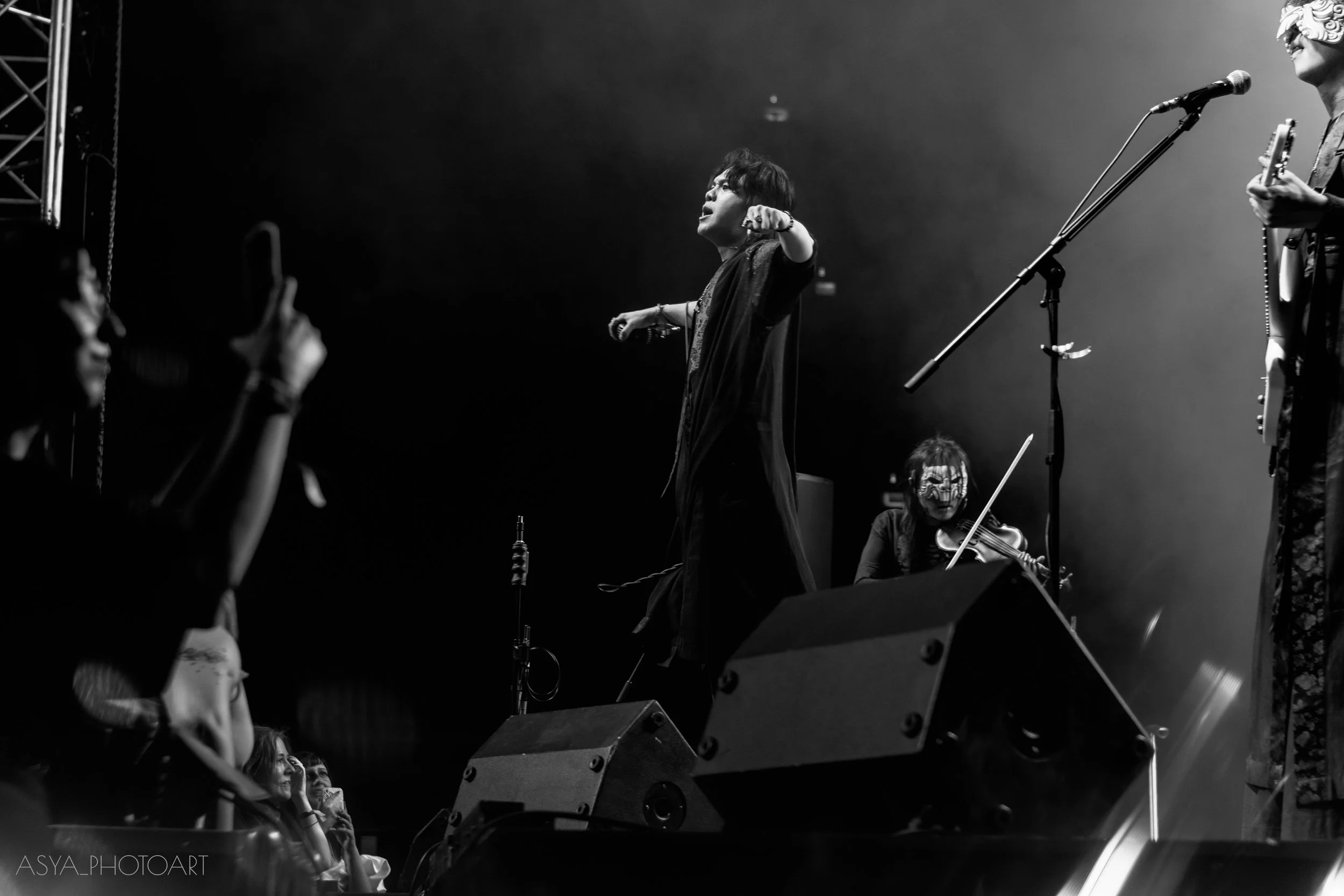 Black and white photo of a singer on stage with microphone, surrounded by audience and musicians, some audience members are taking photos, stage lighting emphasizes the singer and musicians.