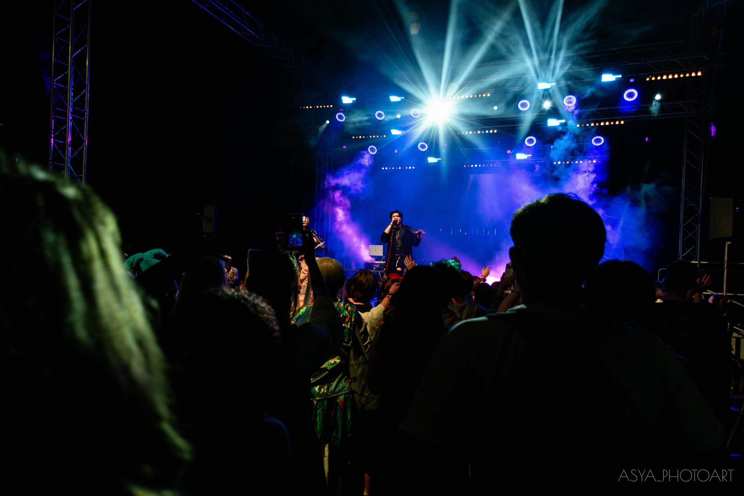 A concert stage illuminated with bright blue and white lights, with a performer singing into a microphone and an audience watching.