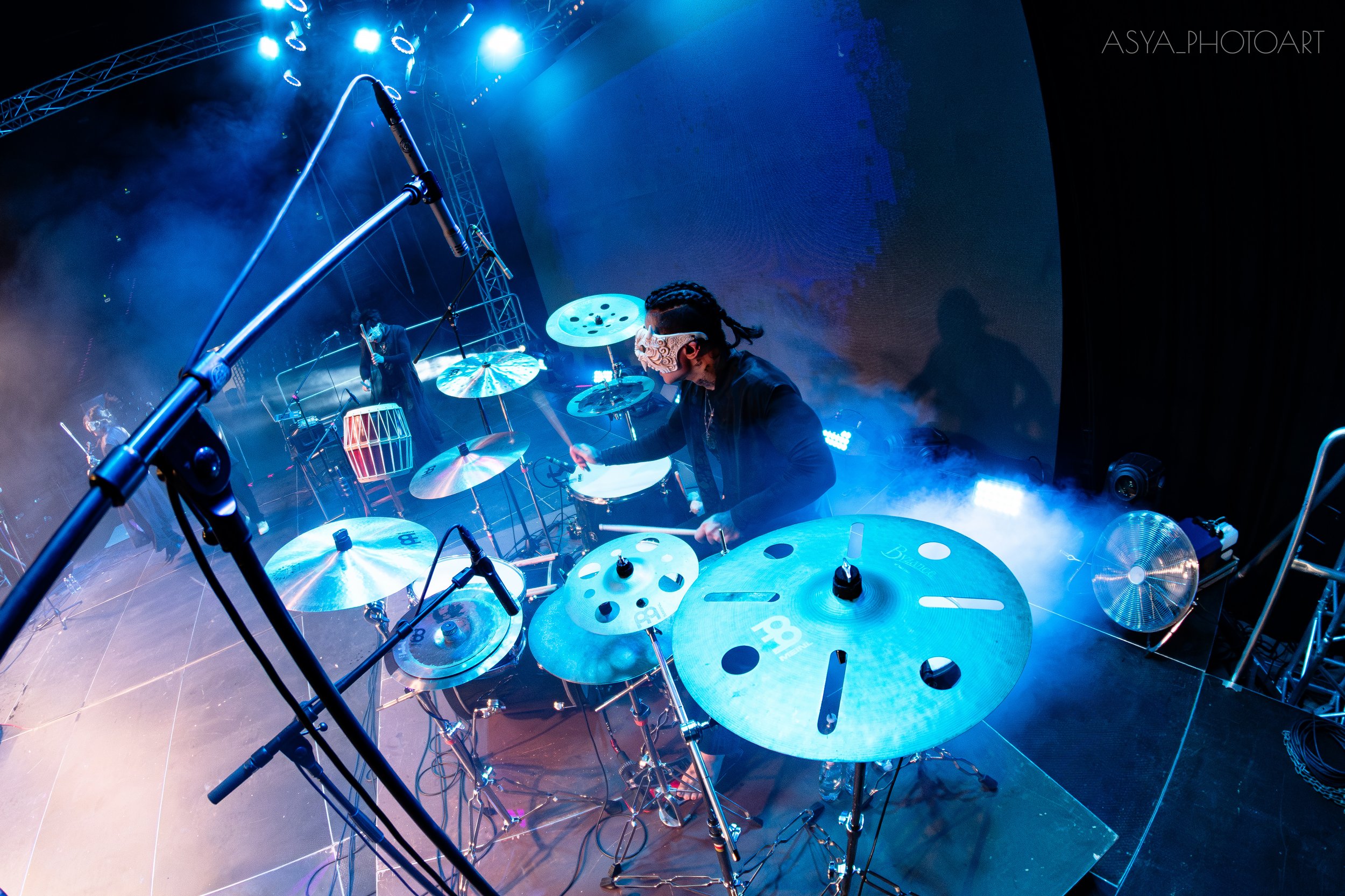 A drummer playing percussion instruments on a stage with blue lighting, surrounded by microphones and stage equipment.