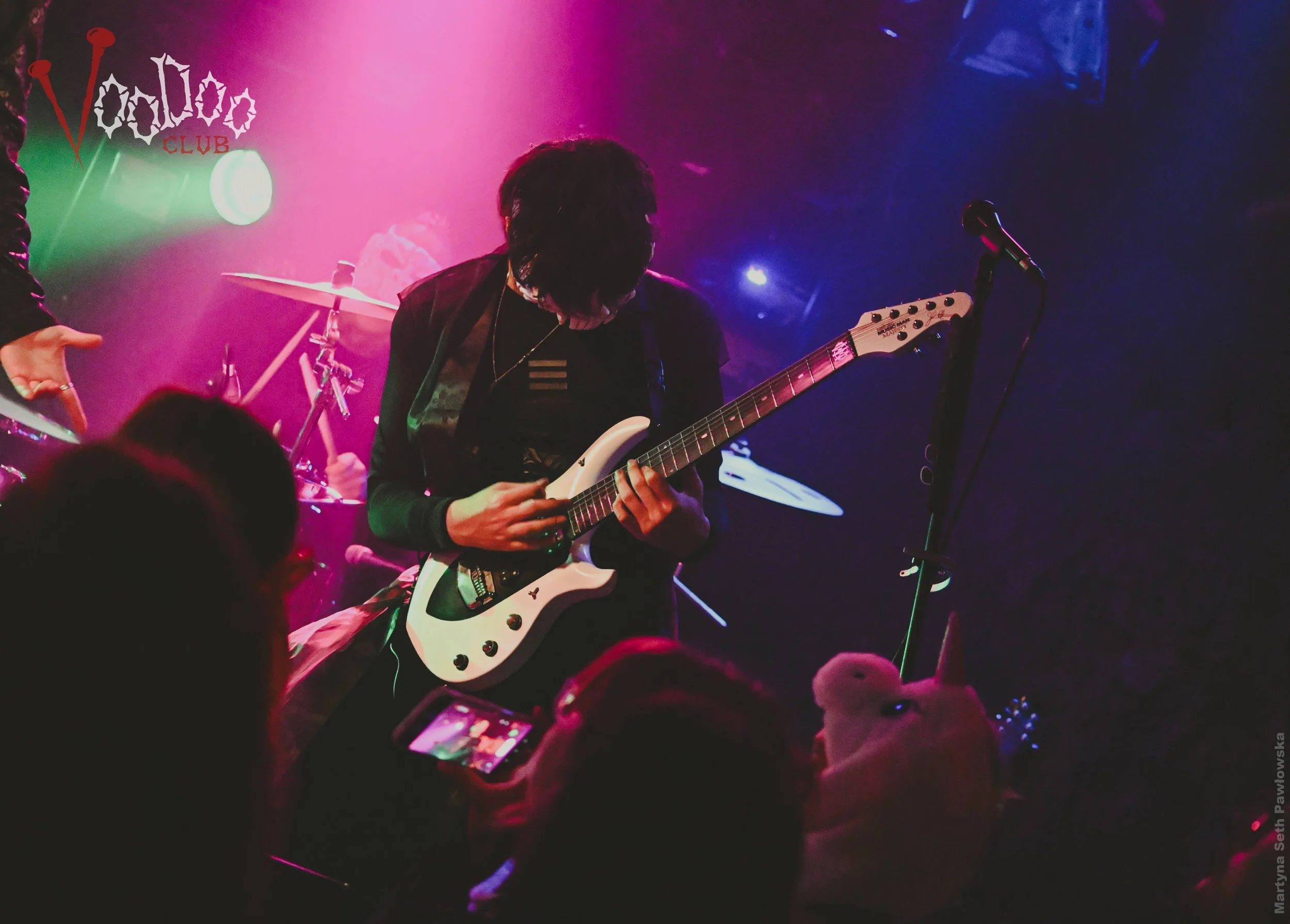 A musician playing an electric guitar on stage with colorful lighting. The audience is visible in the foreground, and there is a microphone stand nearby. A sign in the background says 'Voodoo Club'.