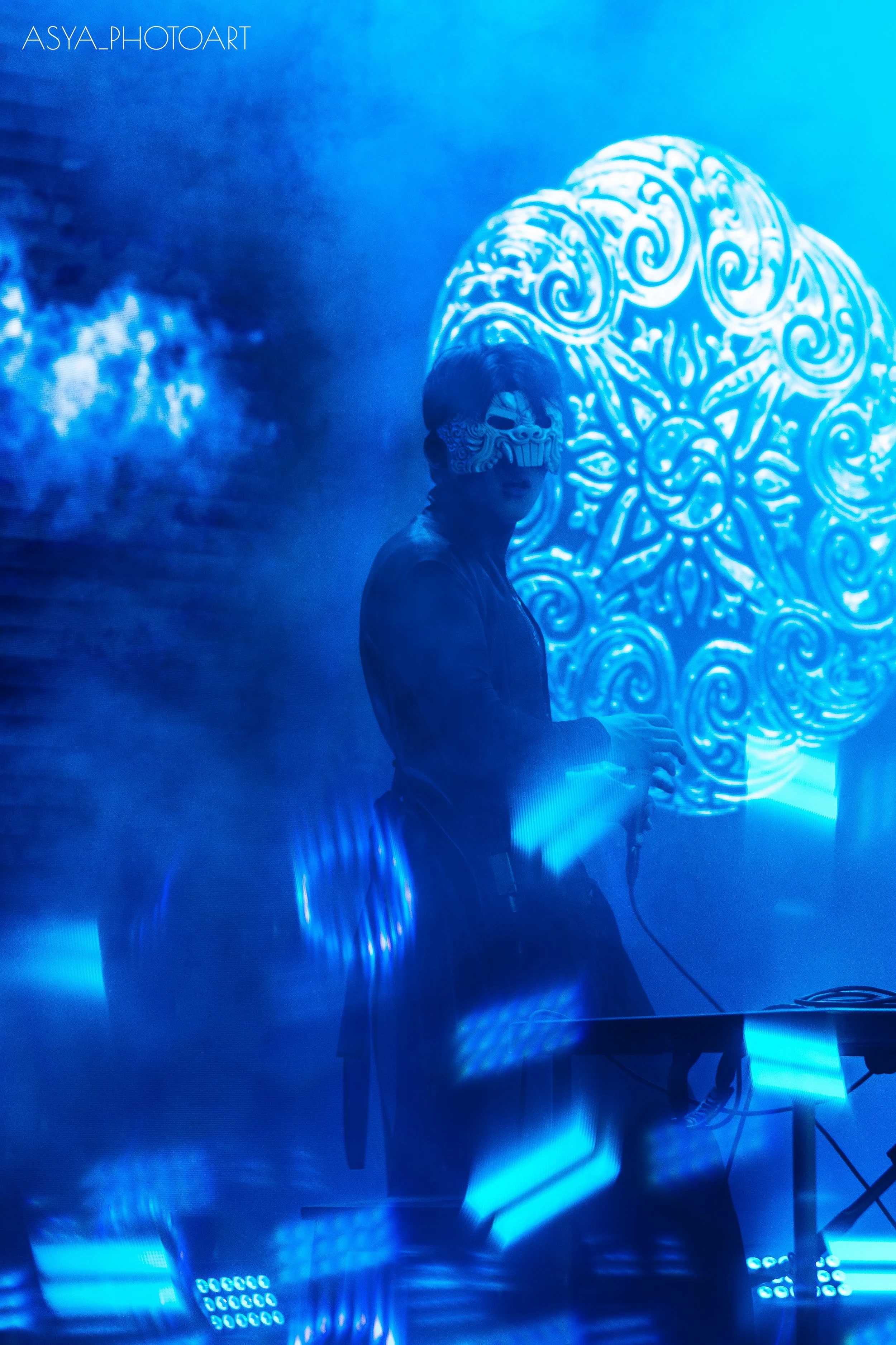 Person wearing a luchador mask standing in front of a brightly lit, ornate, blue and white patterned background with blue lighting effects.