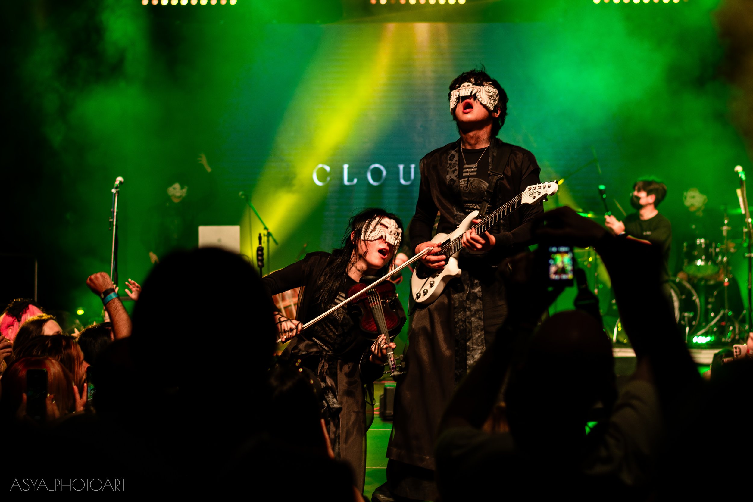 Musicians performing on stage with green and yellow lighting, one guitarist wearing a decorative mask, and other musicians playing instruments.