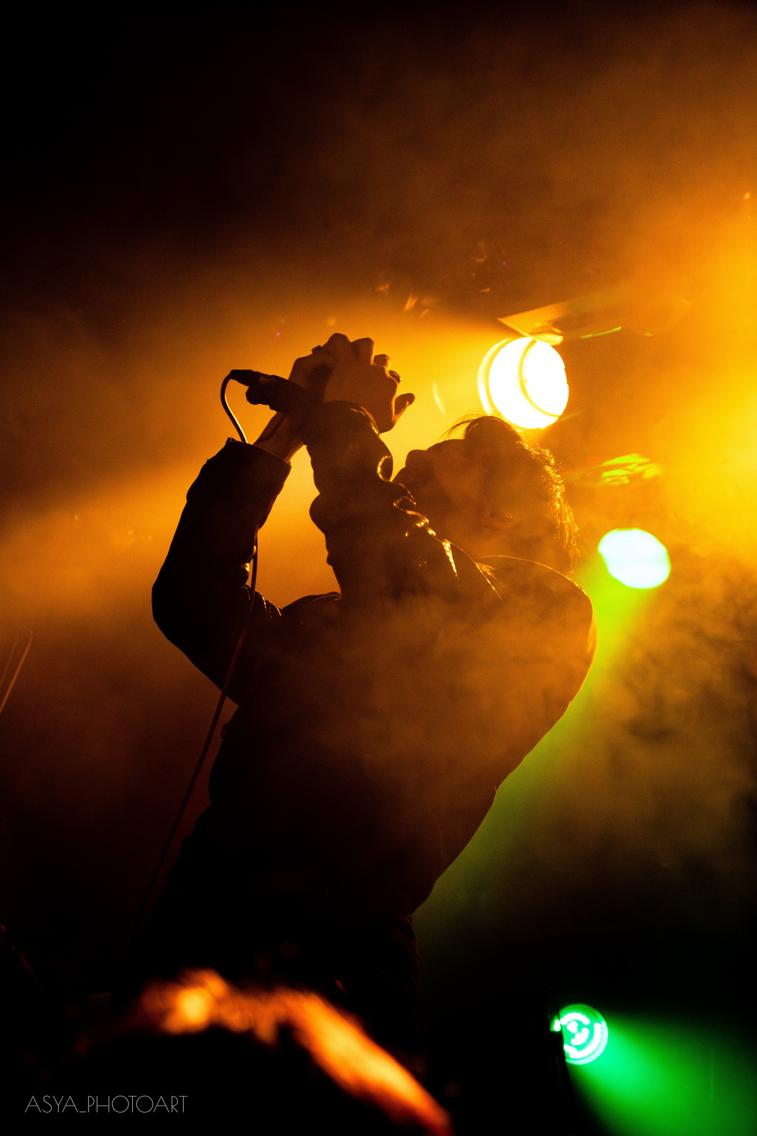 A person singing into a microphone on stage illuminated by yellow stage lights, with a bright yellow and green light in the background.