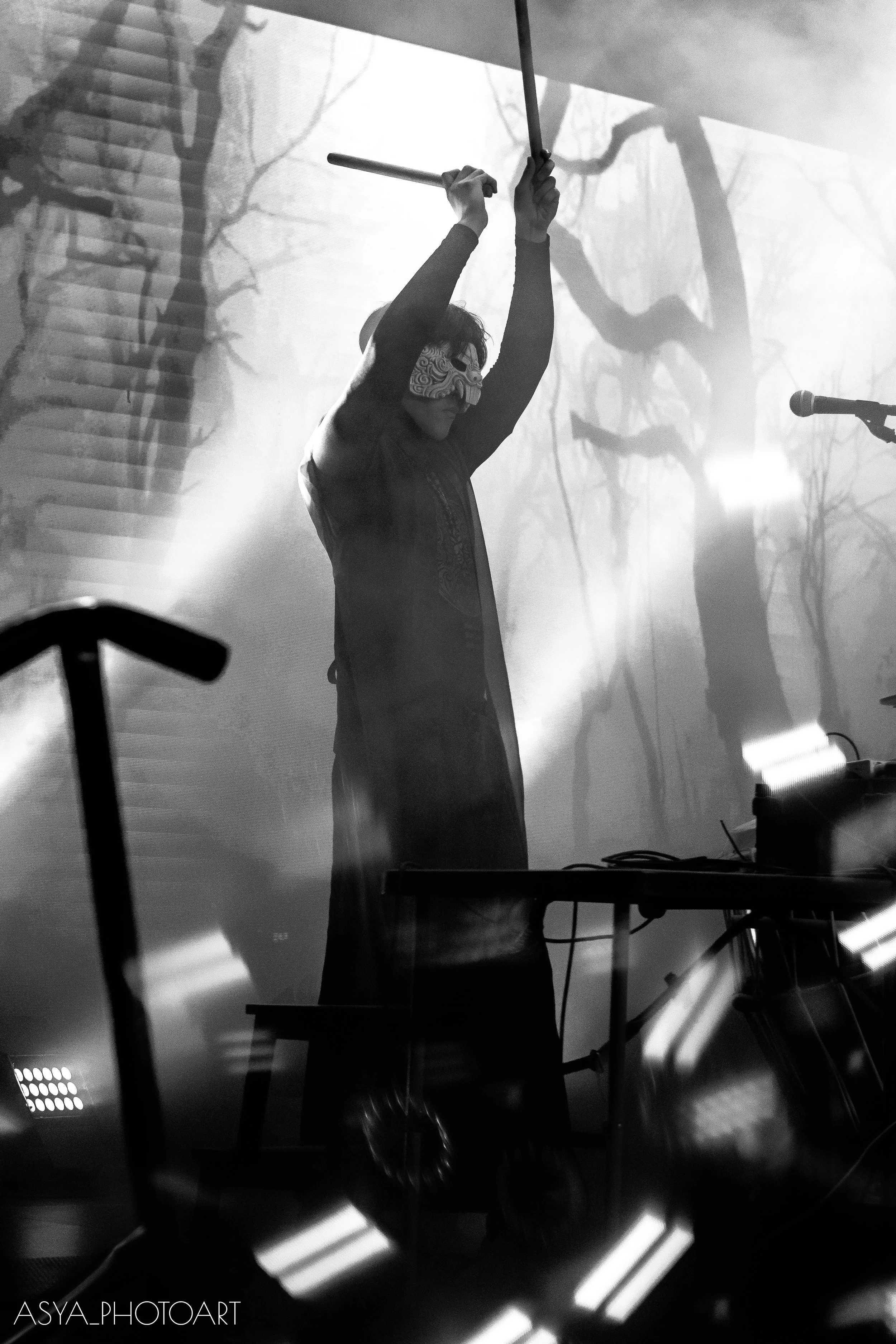 A person on stage wearing a Venetian mask, holding drumsticks raised above their head, with a projection of a face with detailed eye makeup in the background. The scene has dramatic lighting with smoke effects.