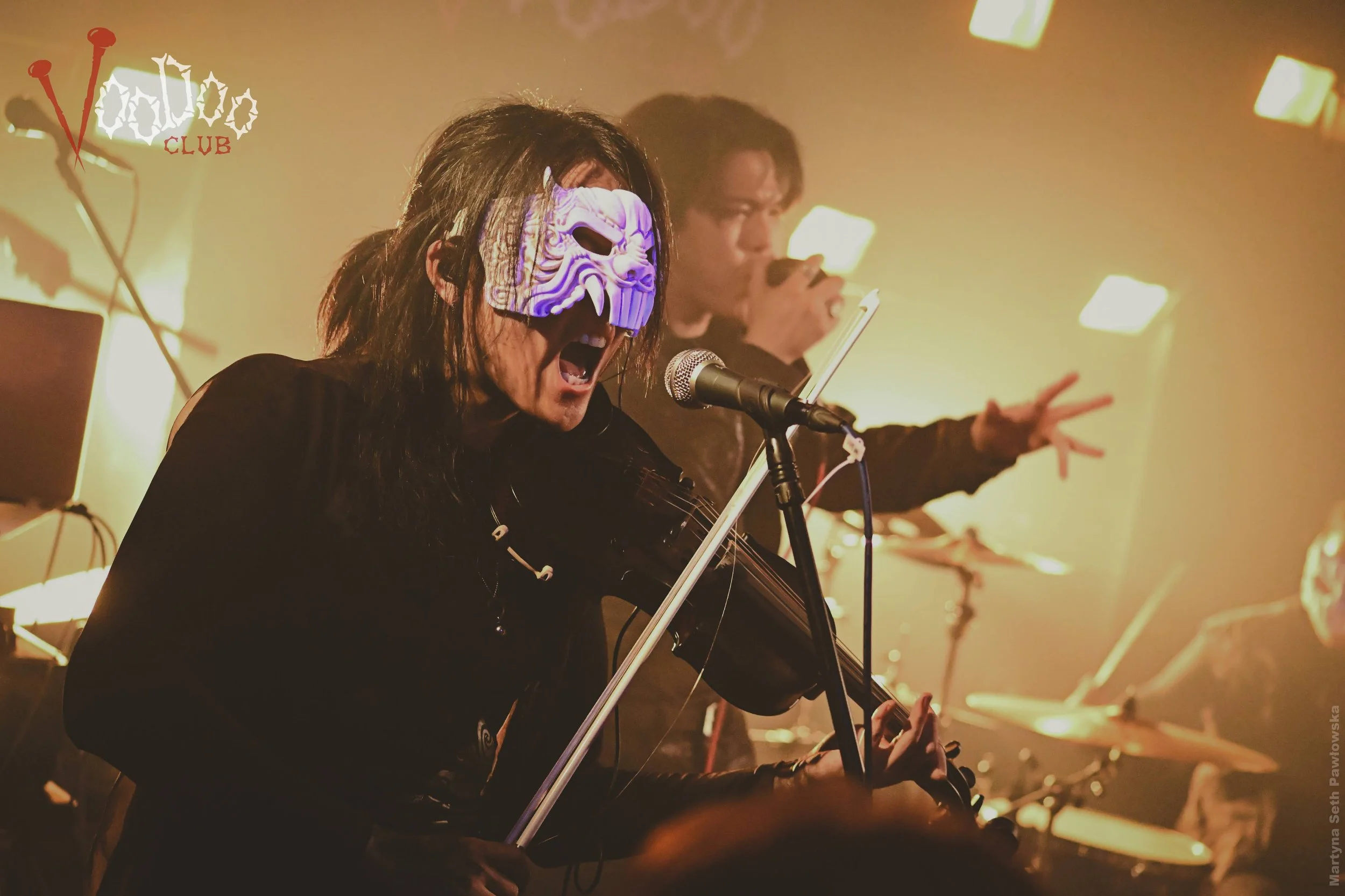 A person wearing a white demon mask with black eyes sings passionately into a microphone while playing a guitar at a live performance, with another person in the background holding a tambourine and gesturing.