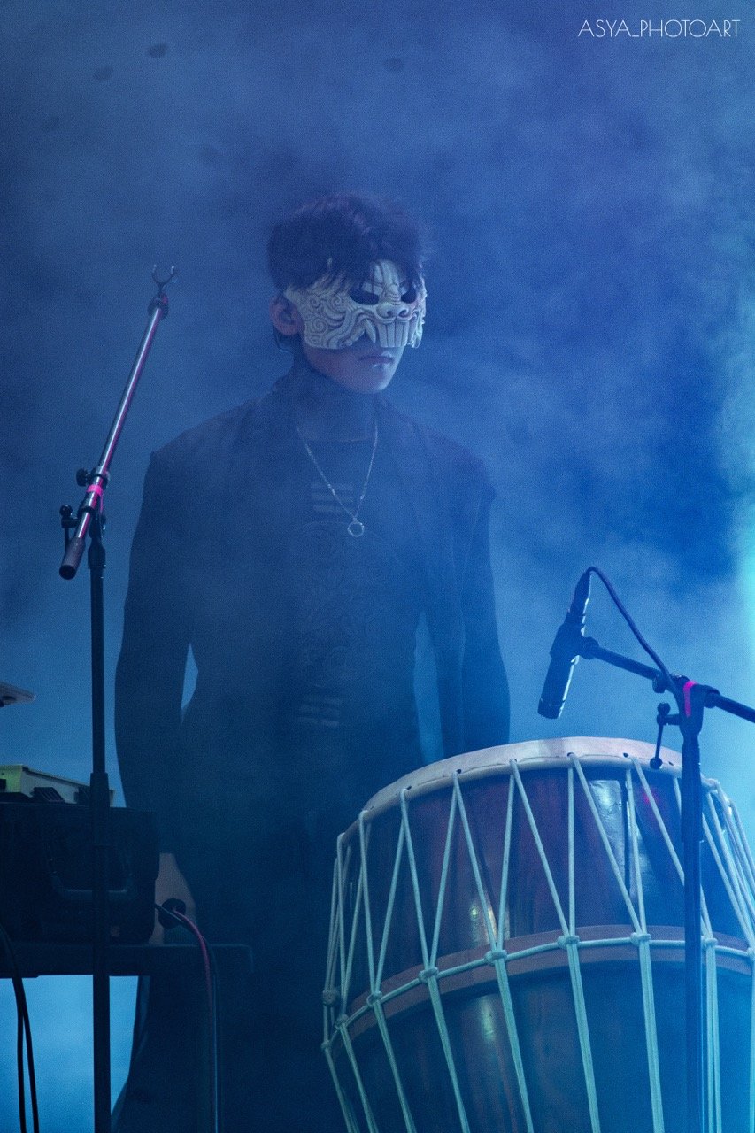 Person wearing a traditional mask playing a drum on stage with microphones and fog.