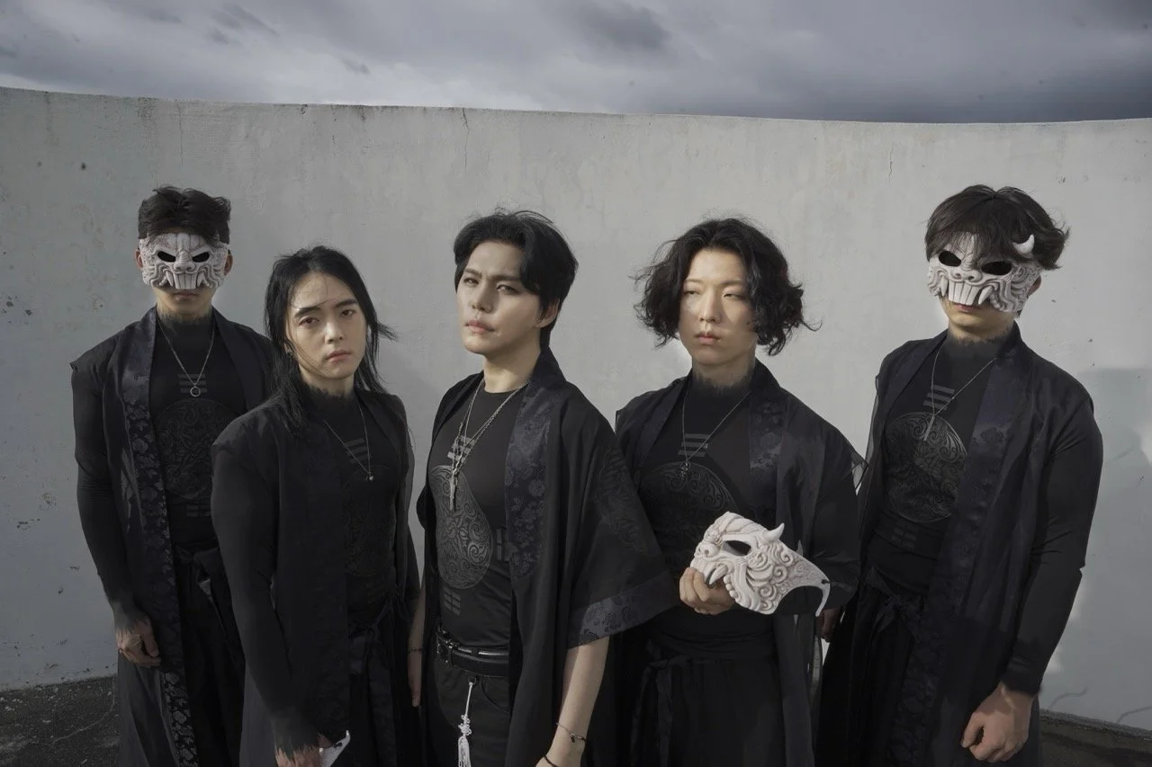 Five people dressed in black clothing, some wearing traditional masks, standing outdoors against a cloudy sky background.
