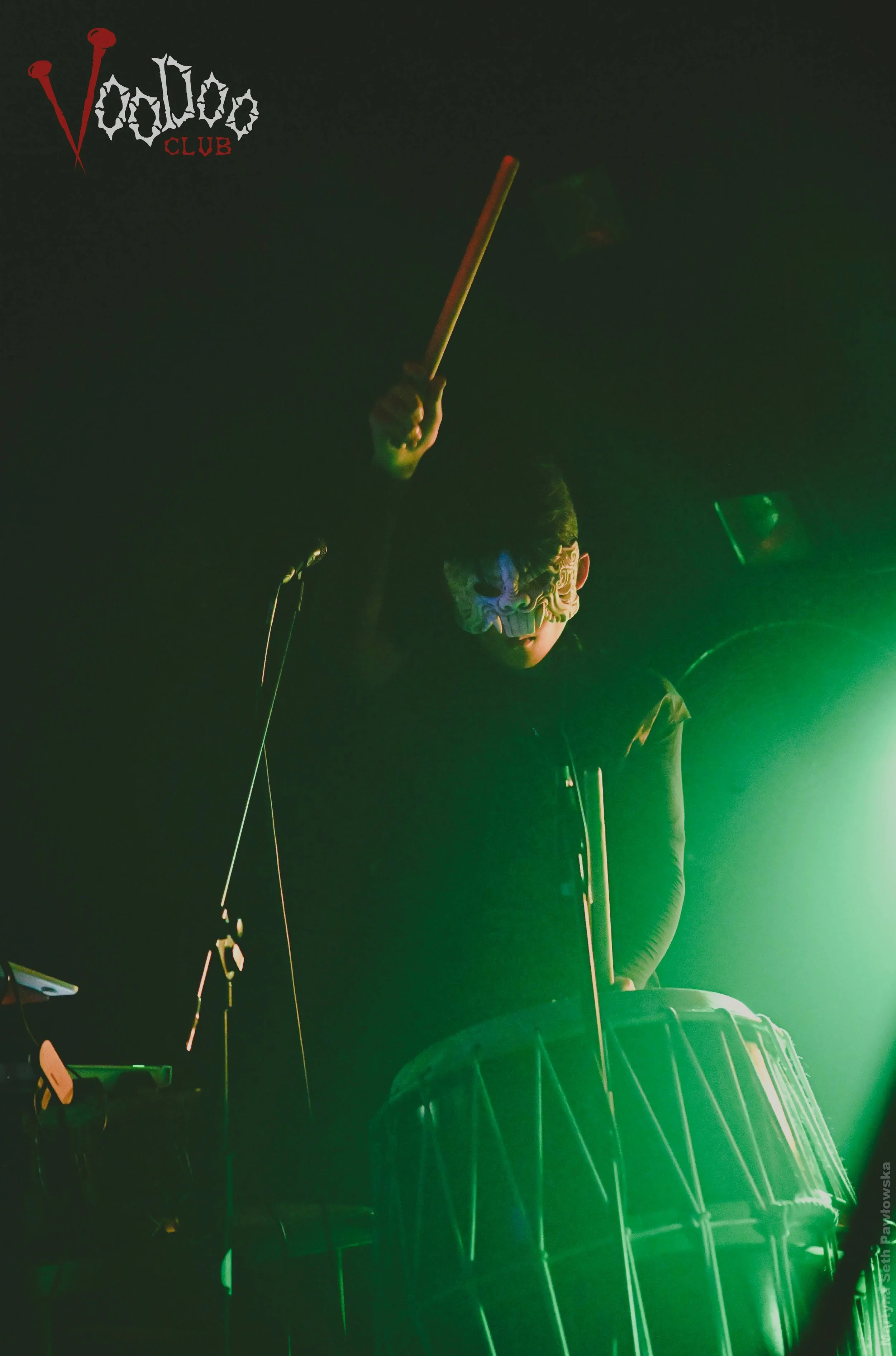 Person wearing a mask with a creepy face design, playing drums in a dark setting illuminated by green light, with a logo for Voodoo Club in the top left corner.