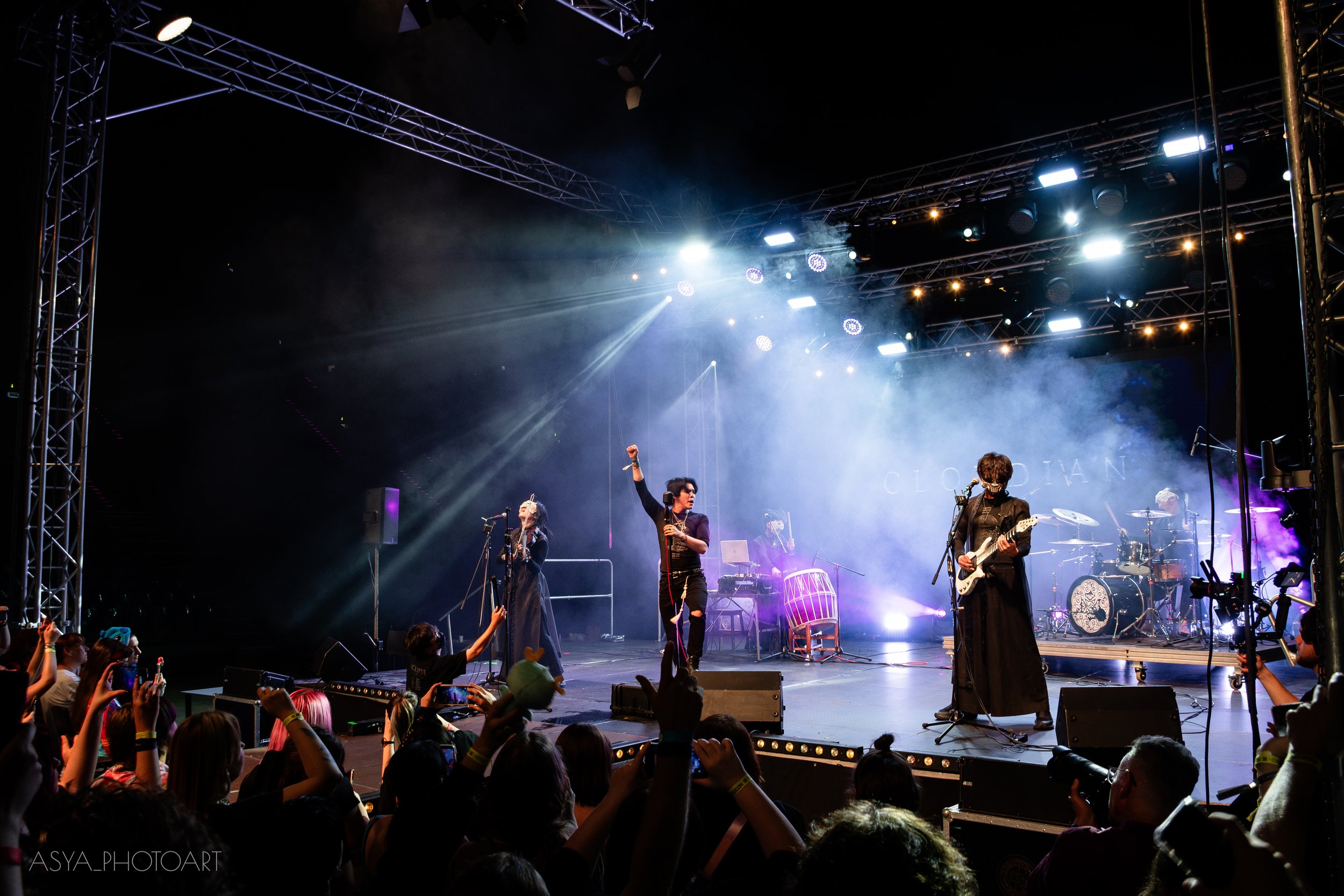 Musicians performing on stage at a concert, with a crowd watching and cheering in front. The stage is illuminated with bright lights and stage equipment is visible.