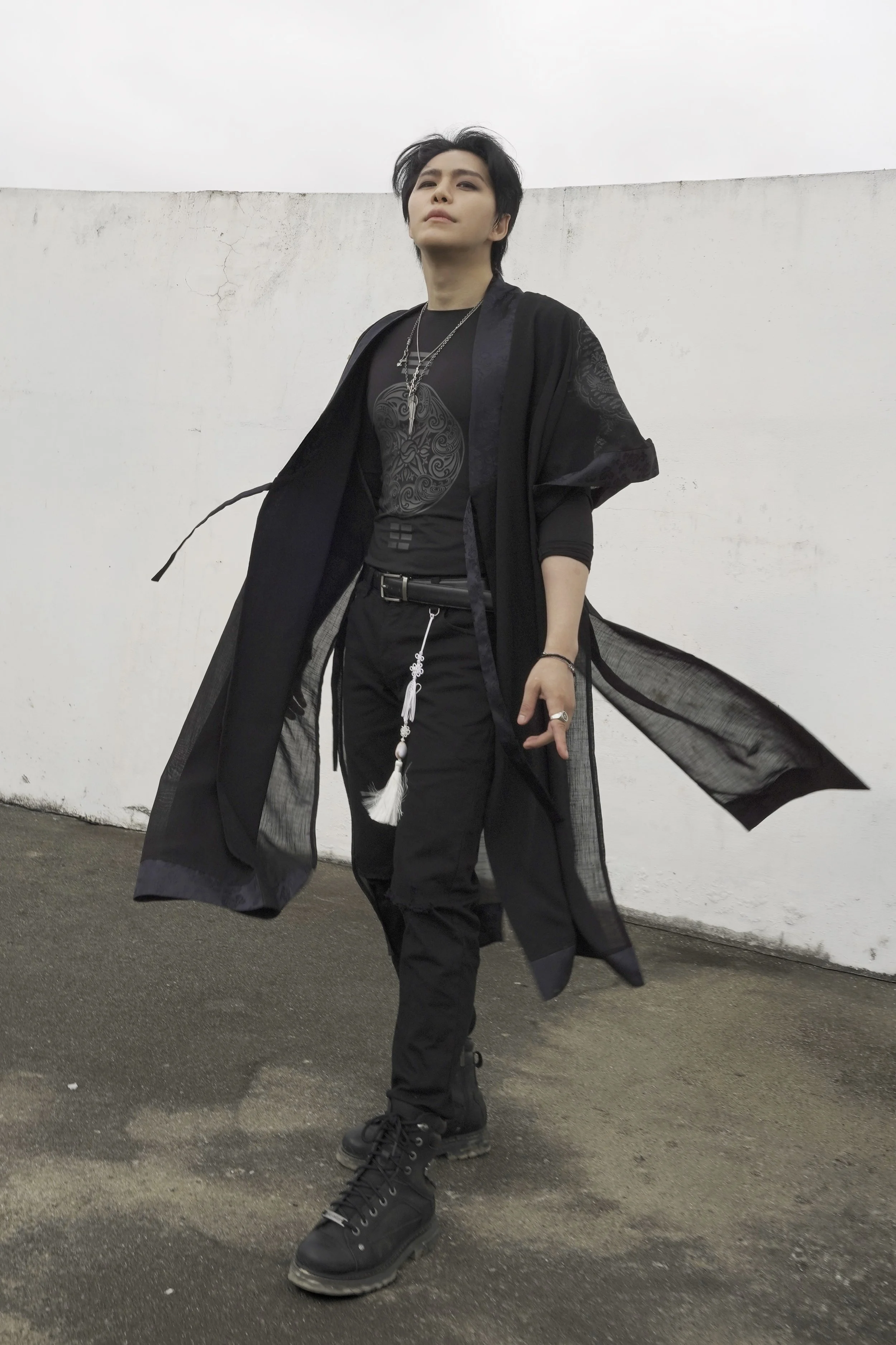 Person standing against a white wall, dressed in black clothing with layered necklaces, wearing black combat boots, with short dark hair, and posing confidently.