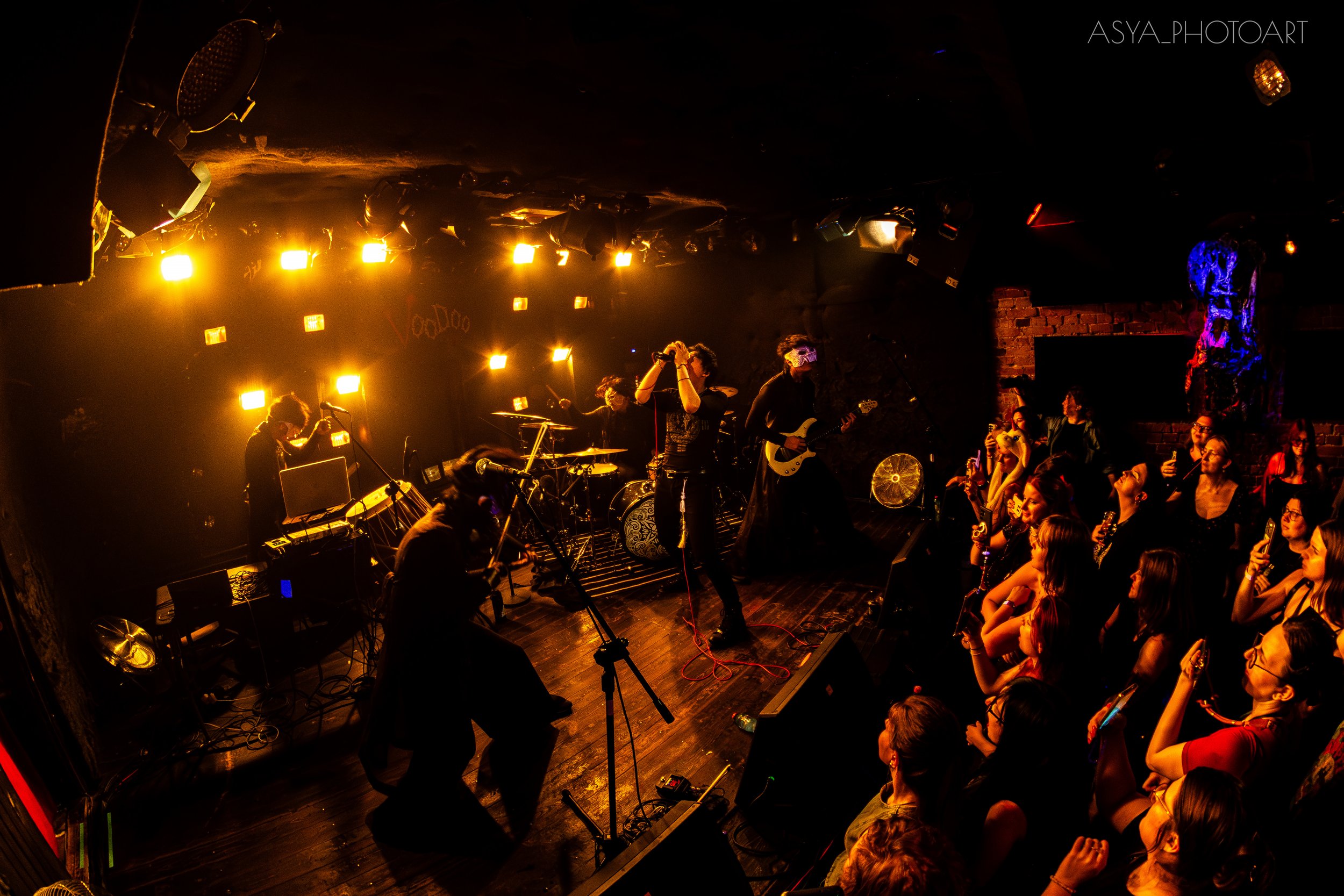 A live musical performance on stage in a dimly lit venue with an audience watching. The band includes a lead singer, drummer, guitarist, and keyboardist. Audience members are standing close to the stage, some taking photos with their phones. The scen