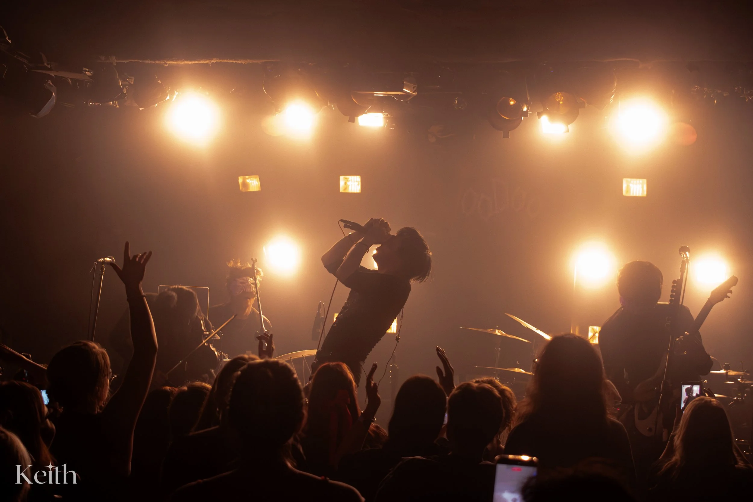 A live music concert with musicians performing on stage, illuminated by bright stage lights, with an audience in front, some raising their hands.
