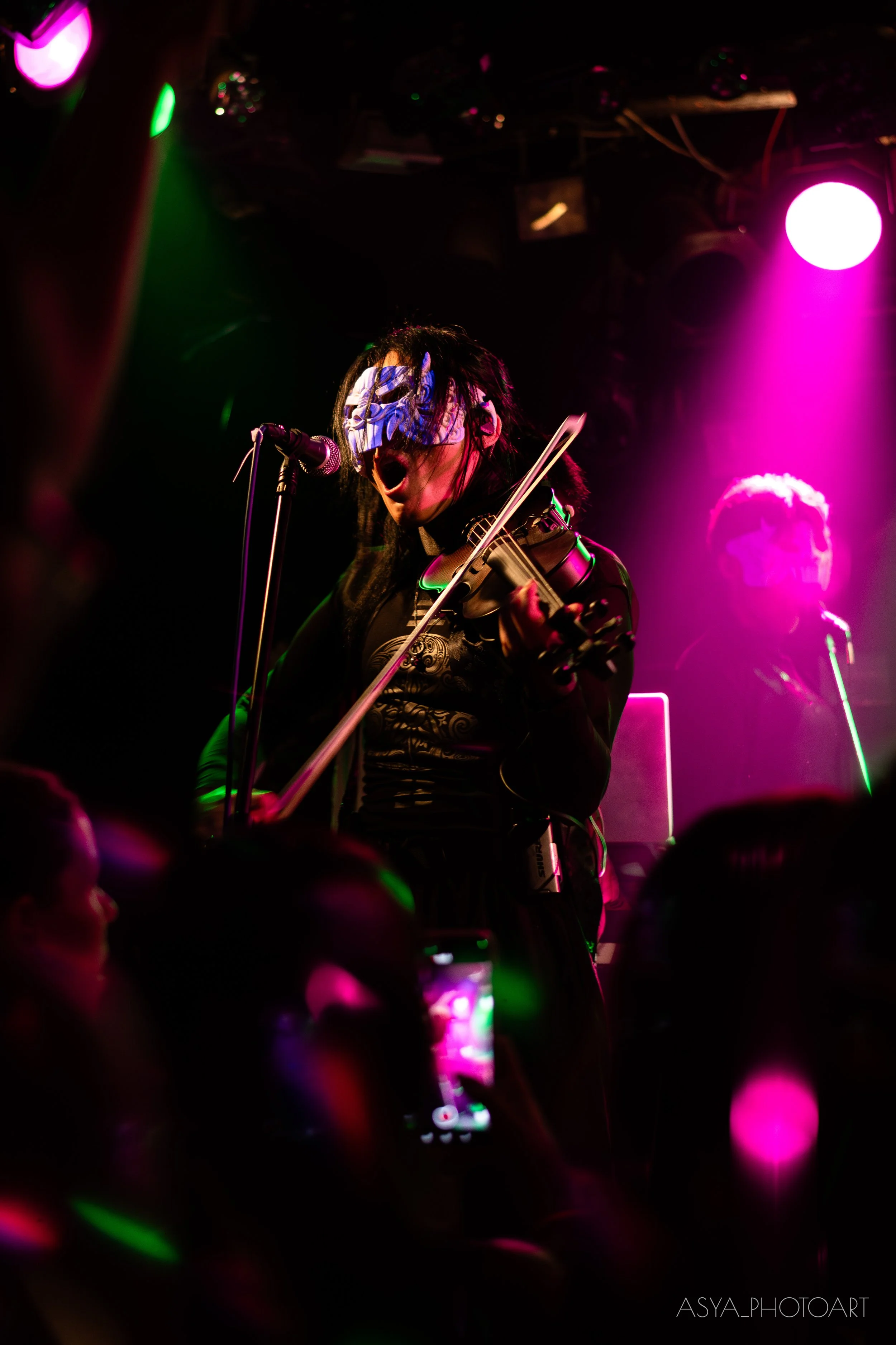 Performer wearing a mask and playing a violin on stage, illuminated by pink and green stage lights with a dark background, audience visible in the foreground.