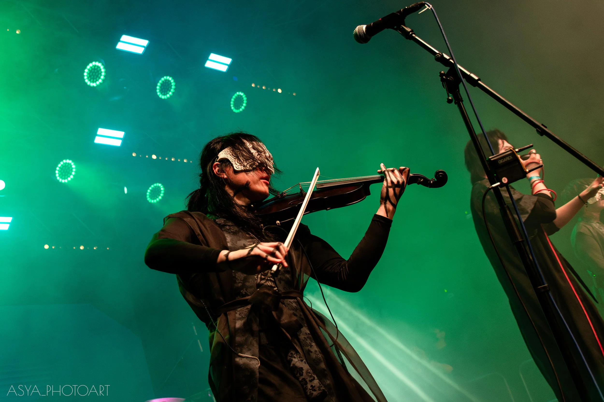 A woman playing the violin on stage, wearing a patterned bandana over her eyes, with a microphone and green background lights.