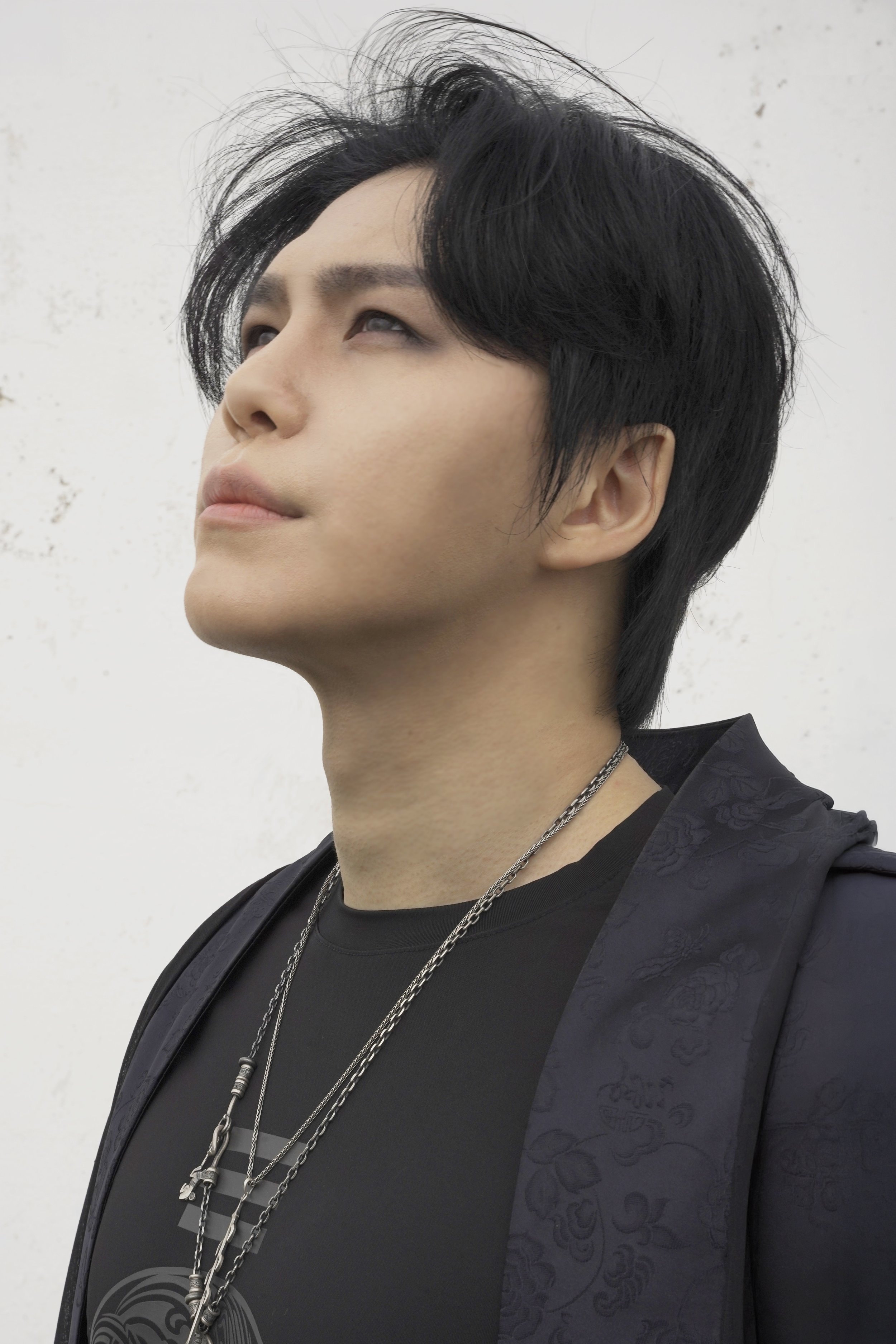 Close-up of a young man with black hair styled to one side, looking upwards, against a plain white background. He is wearing layered necklaces and a black shirt with a dark patterned blazer.