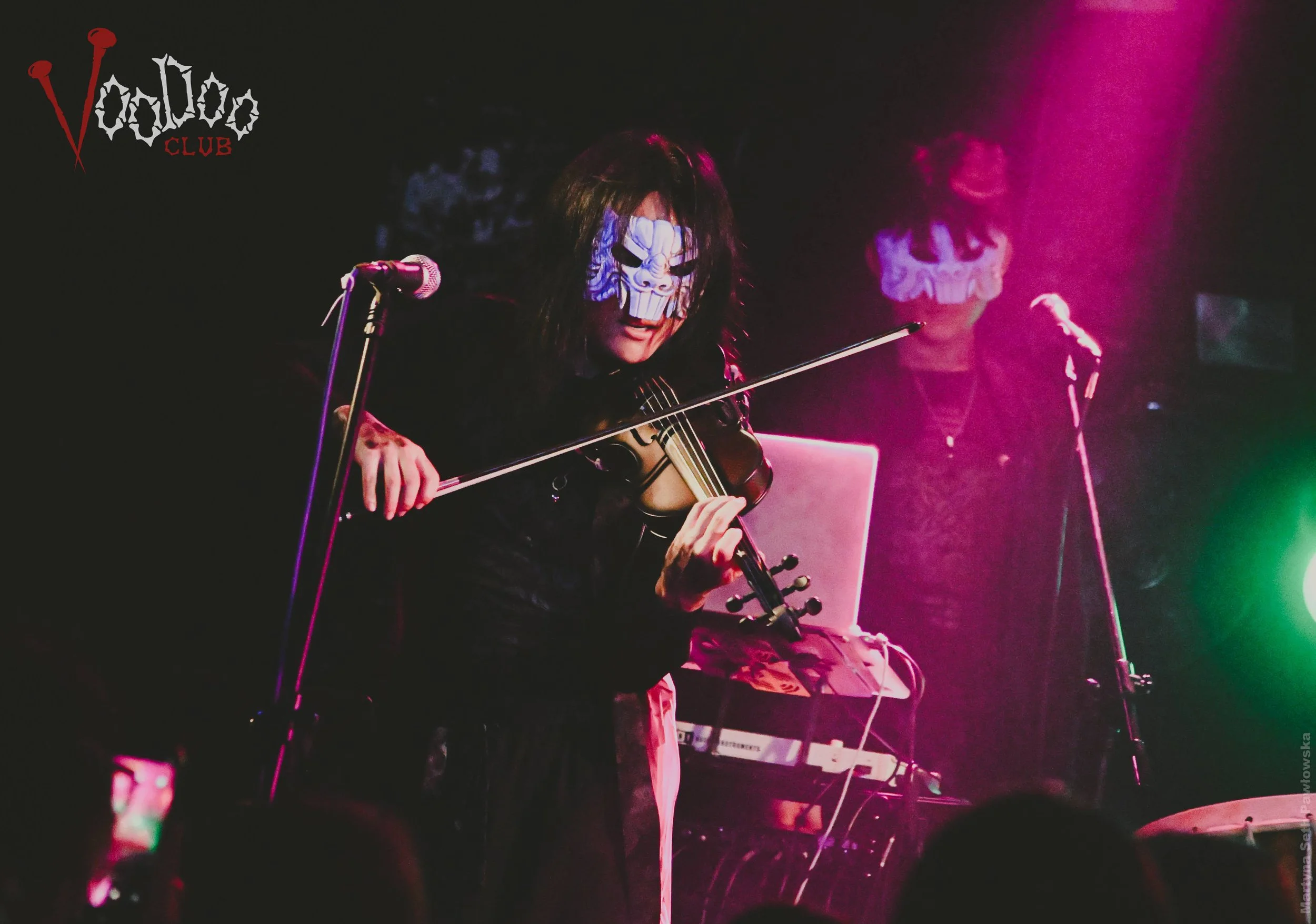 Two musicians wearing skeletal face paint playing instruments on stage with colorful lighting.