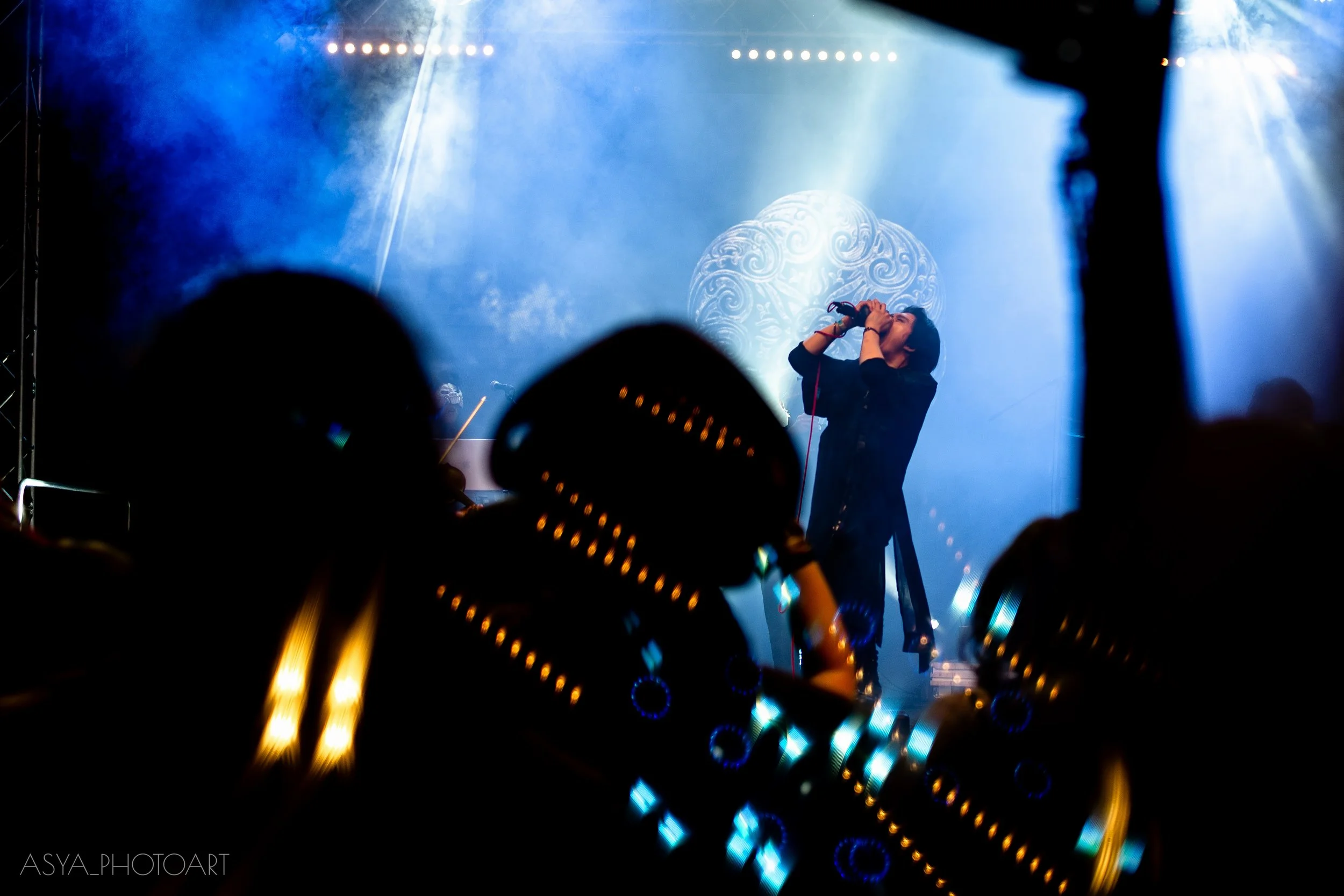 A live concert scene showing a singer holding a microphone, illuminated by stage lights, with a blue background and audience in the foreground.