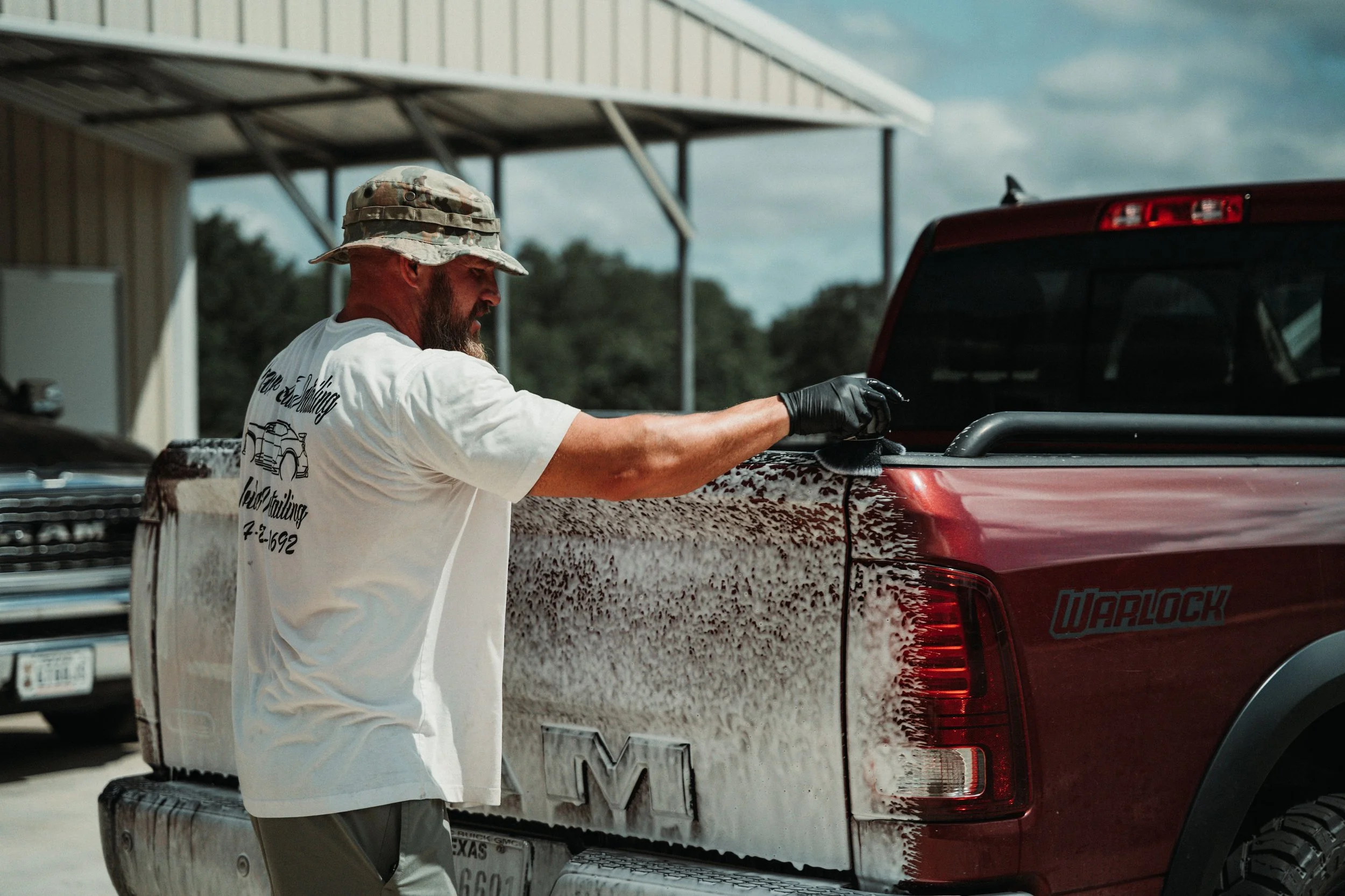 A man with a beard, wearing a camo hat, a white T-shirt, and black gloves, is cleaning foam off the side of a red pickup truck during daytime.