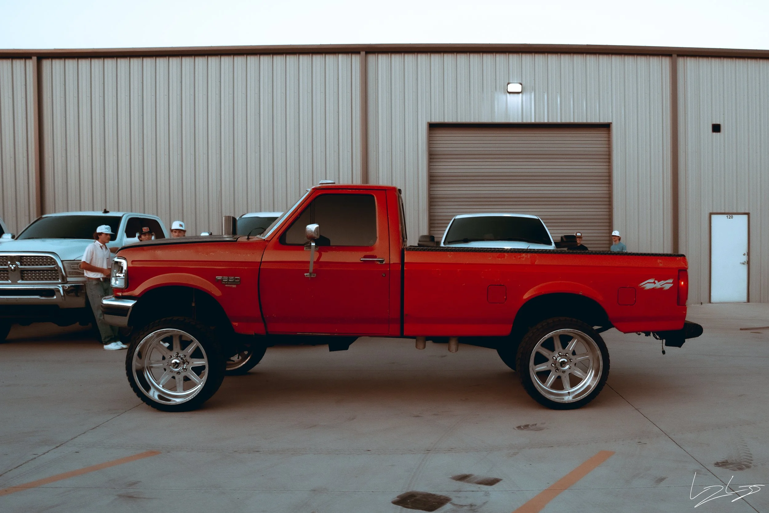 A red pickup truck with large custom wheels parked in front of a warehouse with a beige metal exterior. Several people wearing white baseball caps and casual clothes are standing near other trucks in the background.