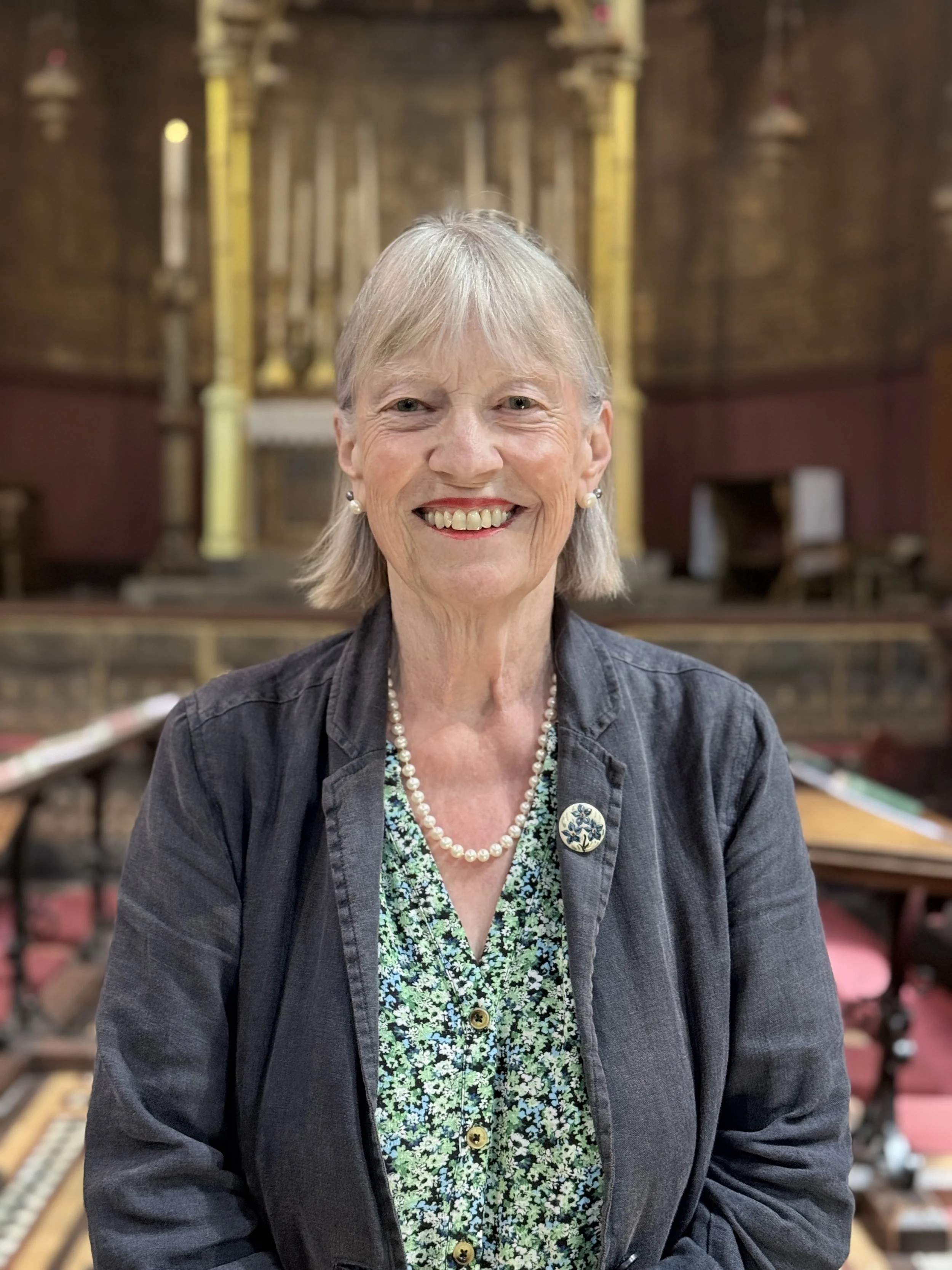 A smiling elderly woman with gray hair, wearing a black jacket, pearl earrings, a pearl necklace, and a floral-patterned dress, standing inside a historic or religious building with wooden pews and a decorative altar in the background.