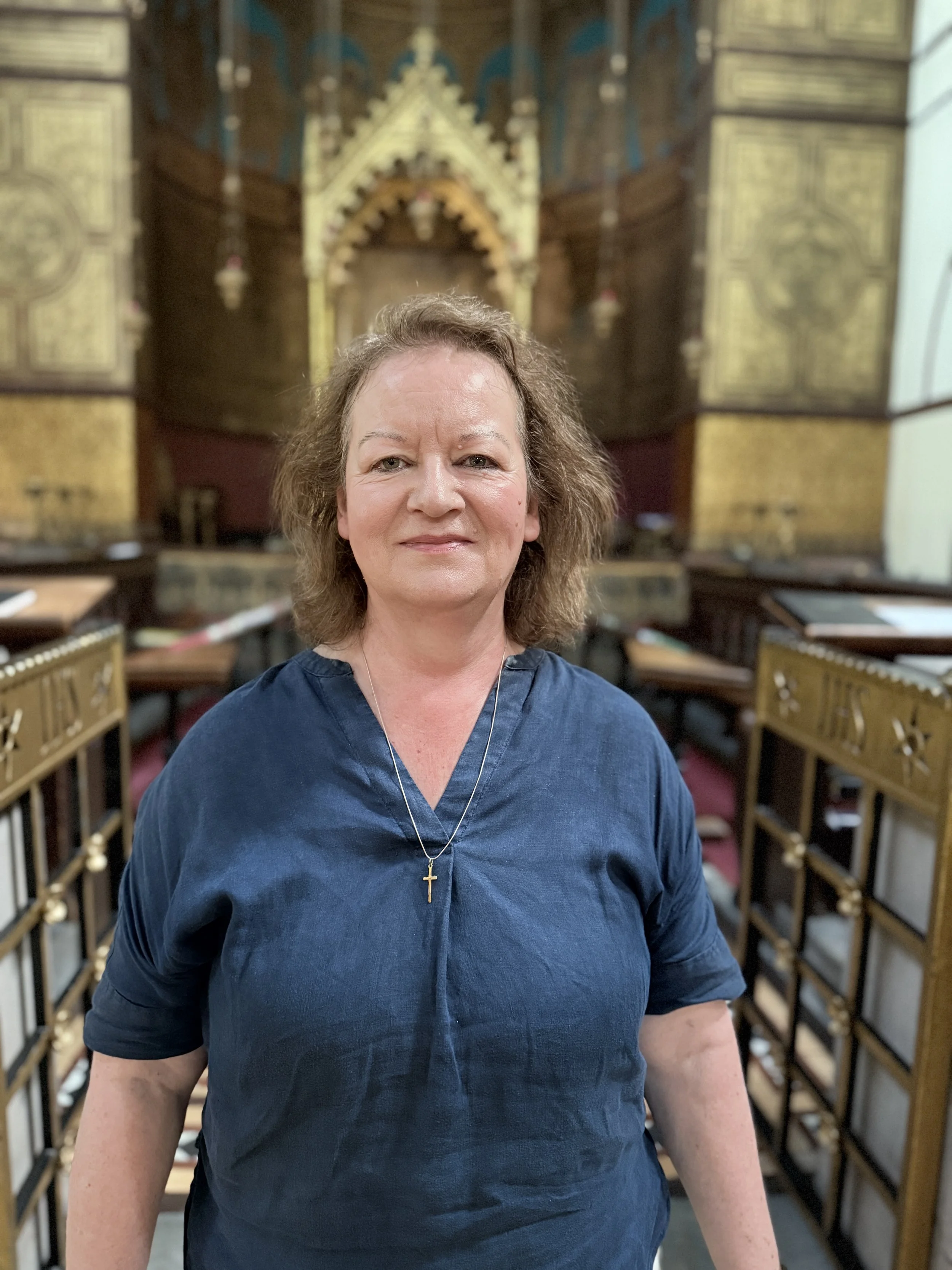 A woman with shoulder-length light brown hair and a cross necklace, wearing a dark blue dress, stands inside a church or chapel with intricate wooden paneling and gold accents in the background.