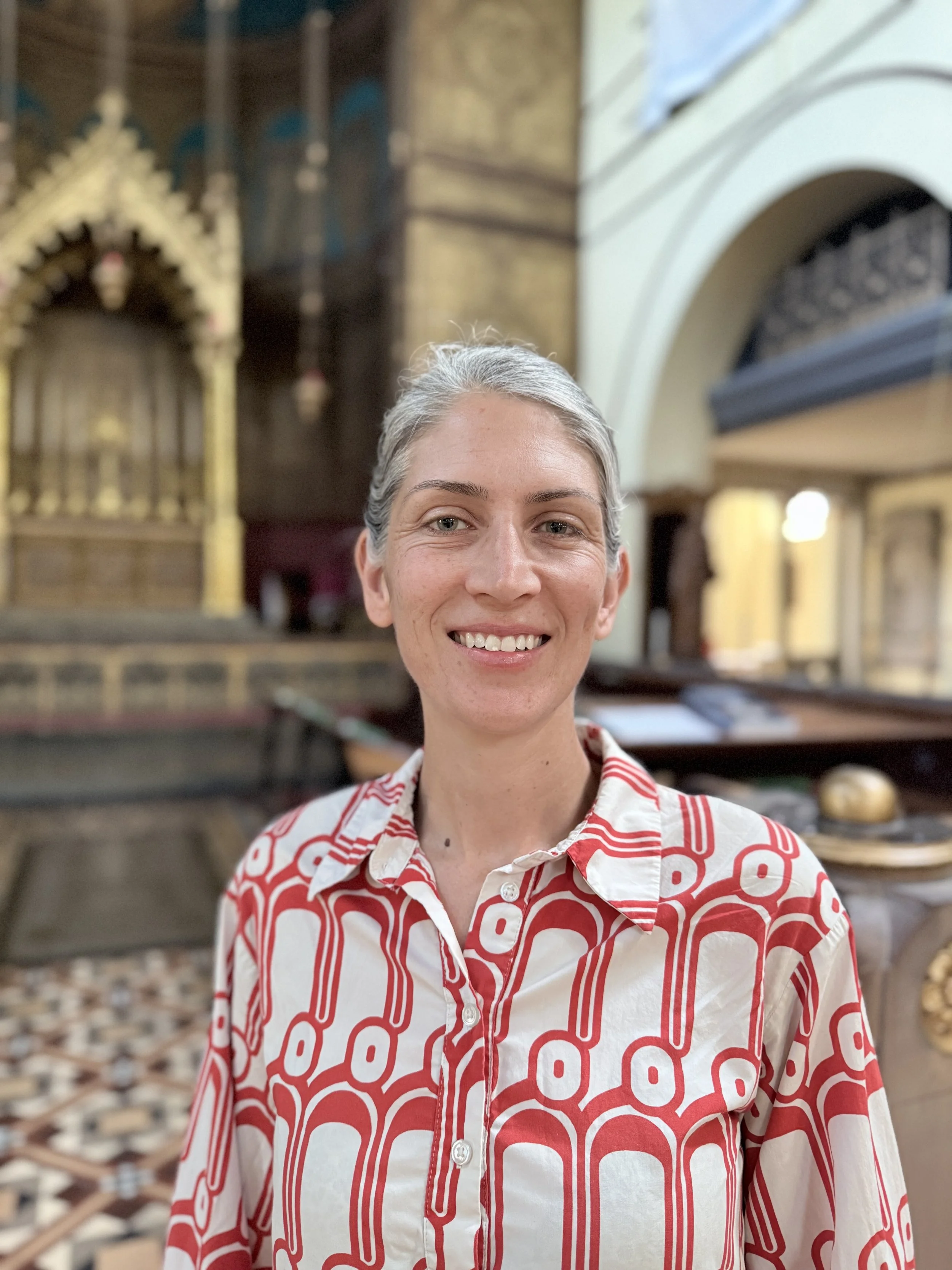 A woman with short gray hair smiling in a church with ornate Catholic altar in the background, wearing a red and white patterned shirt.