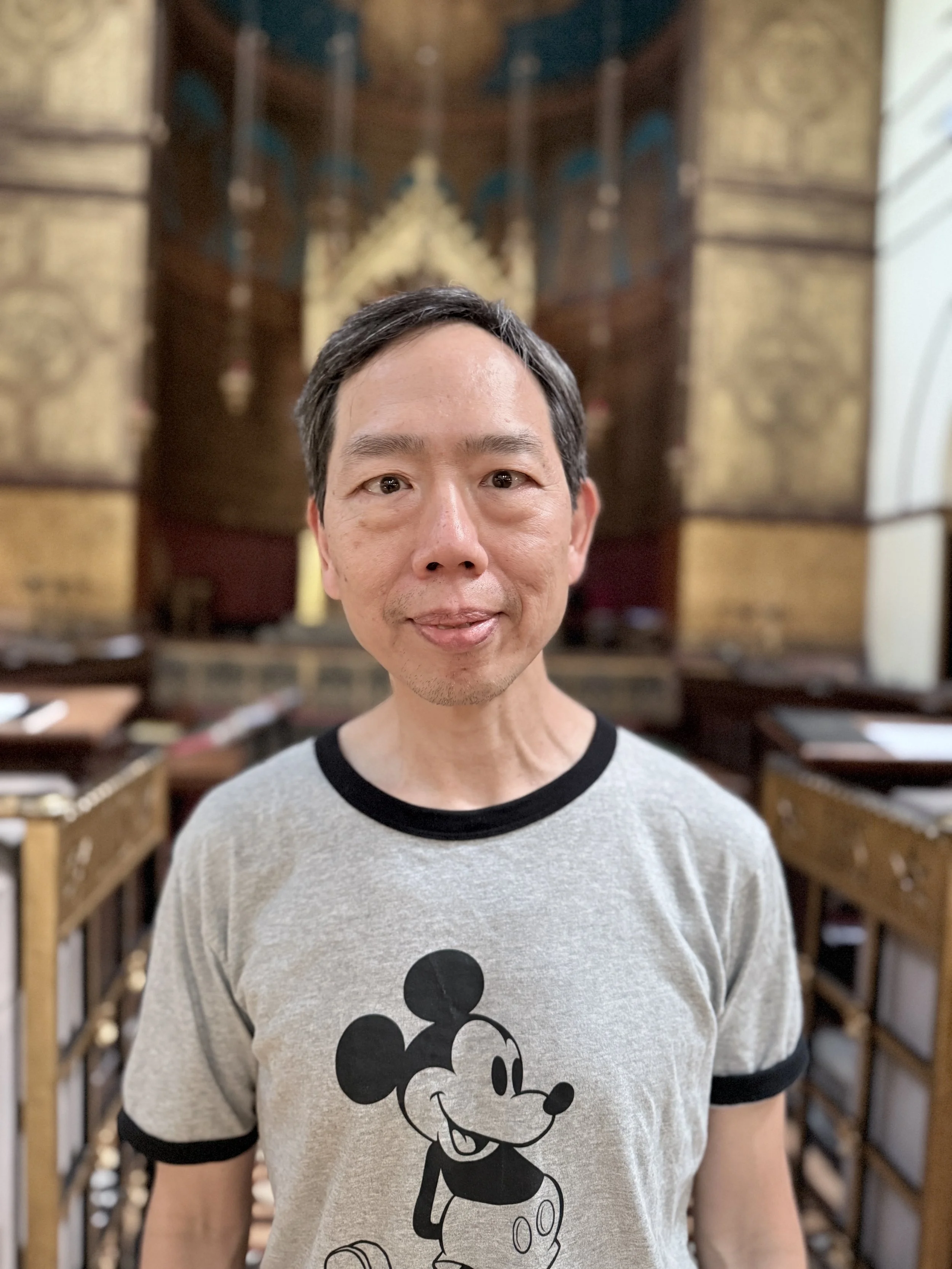 A man with short black hair and a beige Mickey Mouse t-shirt standing in a room with wooden furniture and a decorative wall hanging in the background.