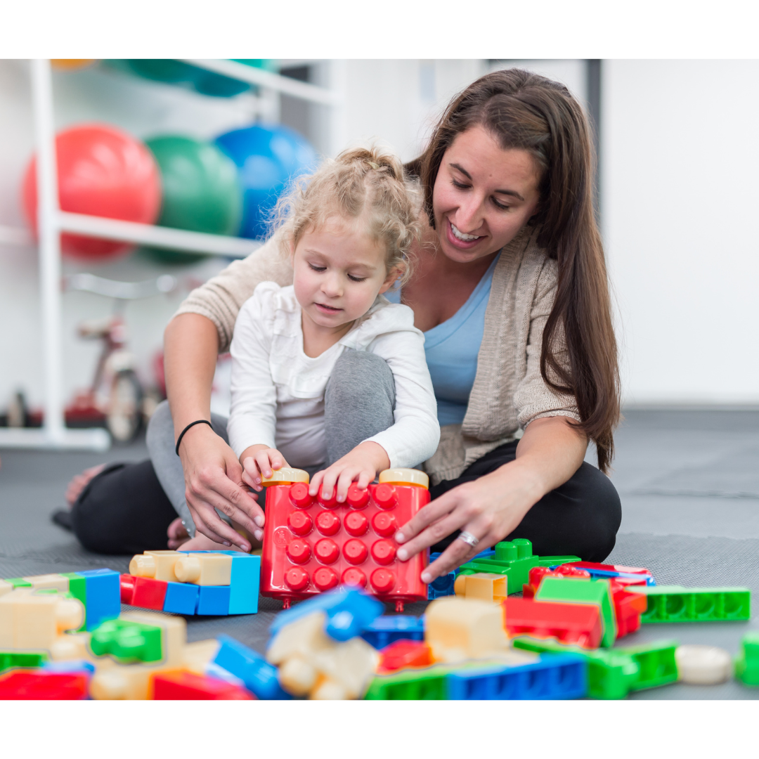 A woman and a young girl playing with colorful building blocks on the floor of a playroom or gym area.