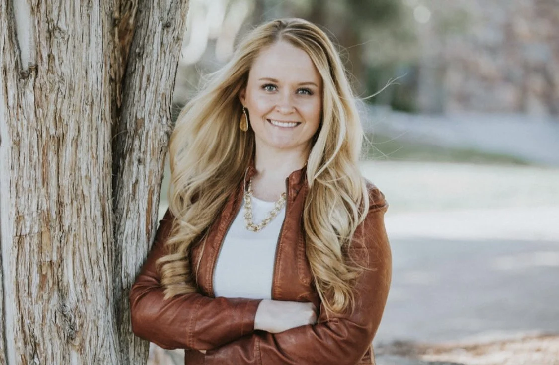 Jennifer Hamelin portrait, smiling, wearing a brown leather jacket and leaning against a tree