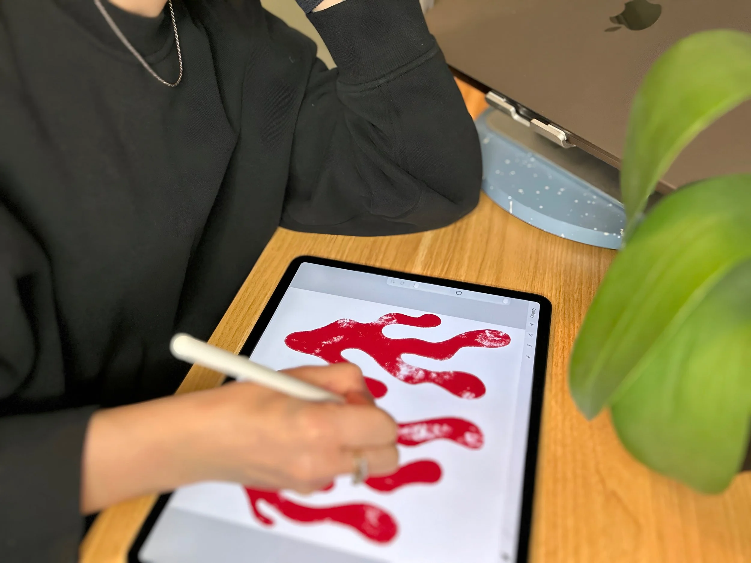 Cat Doxford sketching a red abstract motif on an iPad on a wooden table next to plants and a laptop