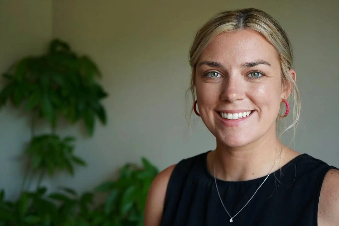 A smiling woman with blonde hair wearing a black sleeveless top, pink hoop earrings, and a delicate necklace, standing indoors with green plants in the background.