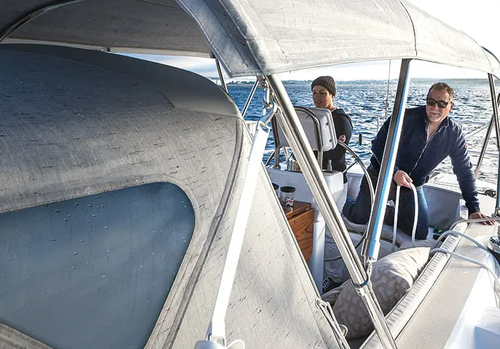 Two people on a boat at sea, with one steering and the other looking over the water, under a canopy.