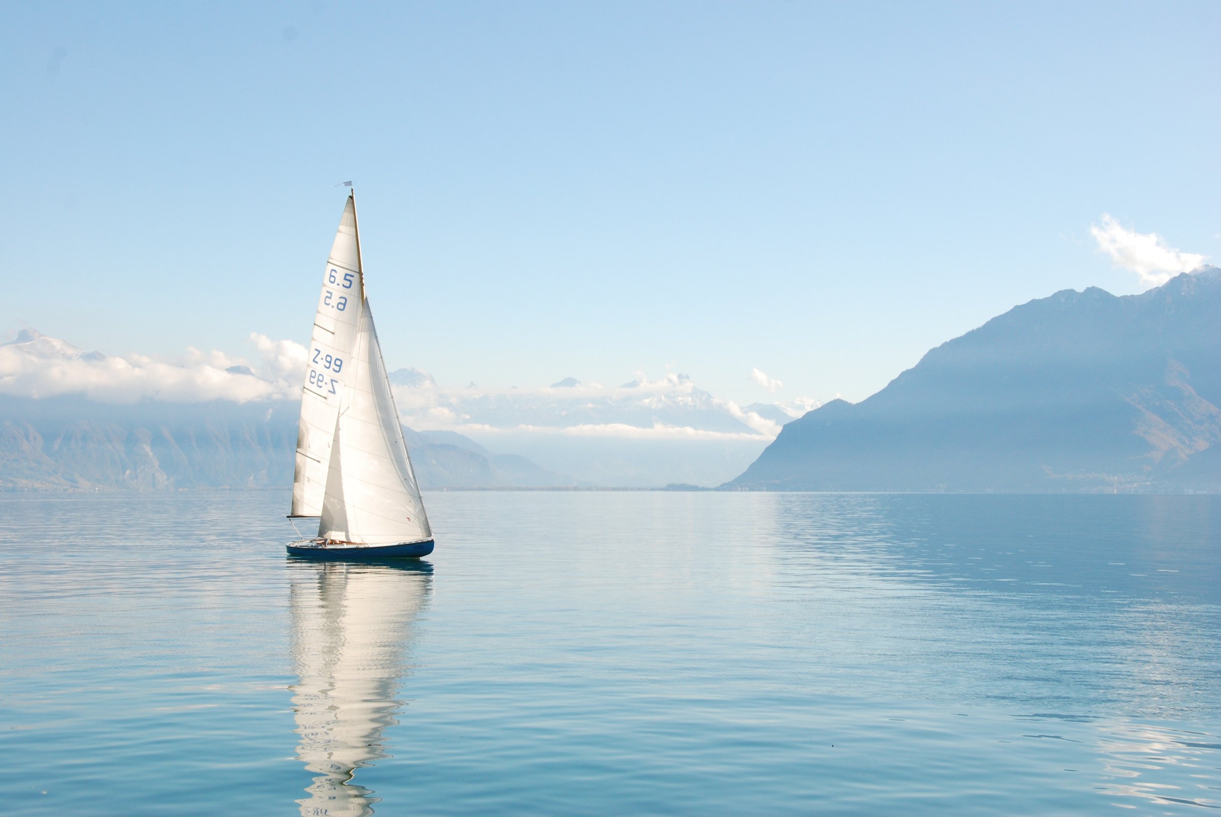 A sailboat with white sails on a calm, reflective body of water, with distant mountains and a partly cloudy sky in the background.