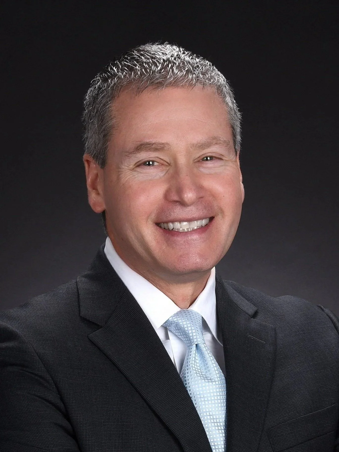 Headshot of a smiling man in a suit and tie against a dark background.