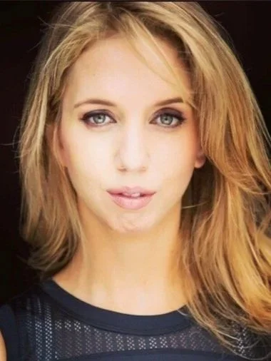 Close-up portrait of a young woman with blonde hair and blue eyes, wearing a dark sleeveless top, against a dark background.