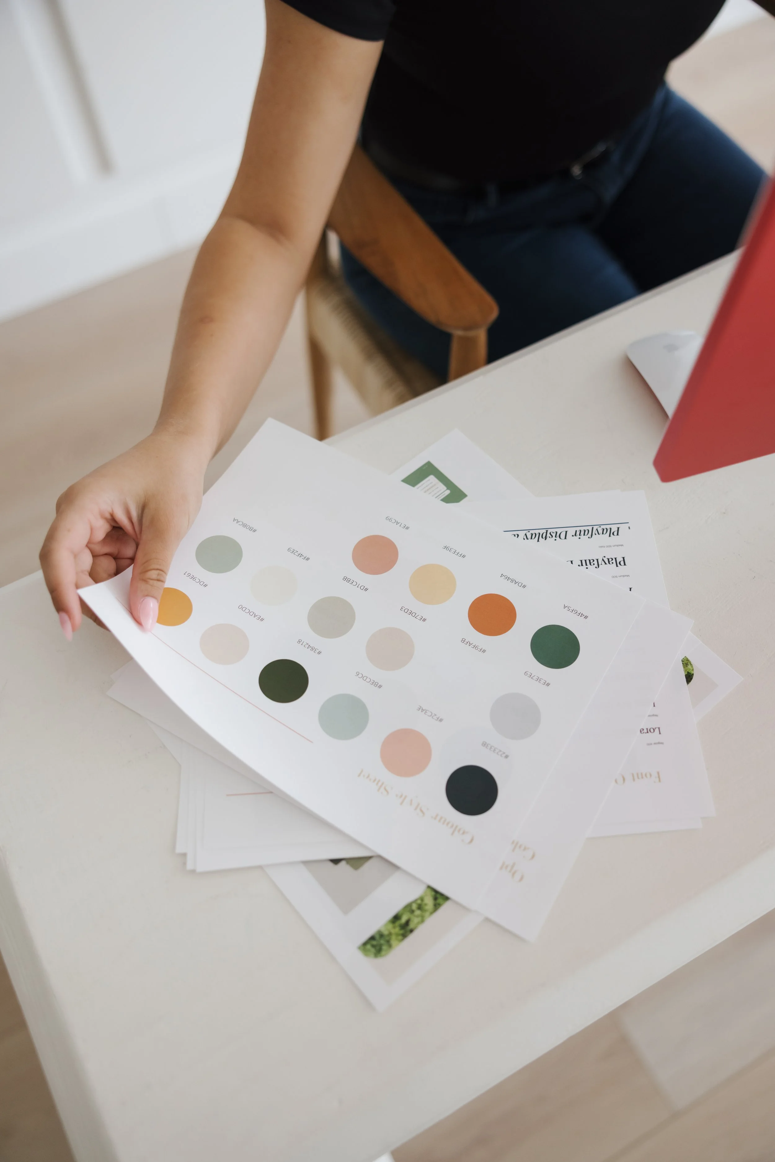 A person holds paper samples with font options and text comparisons at a desk, next to a pink computer monitor.