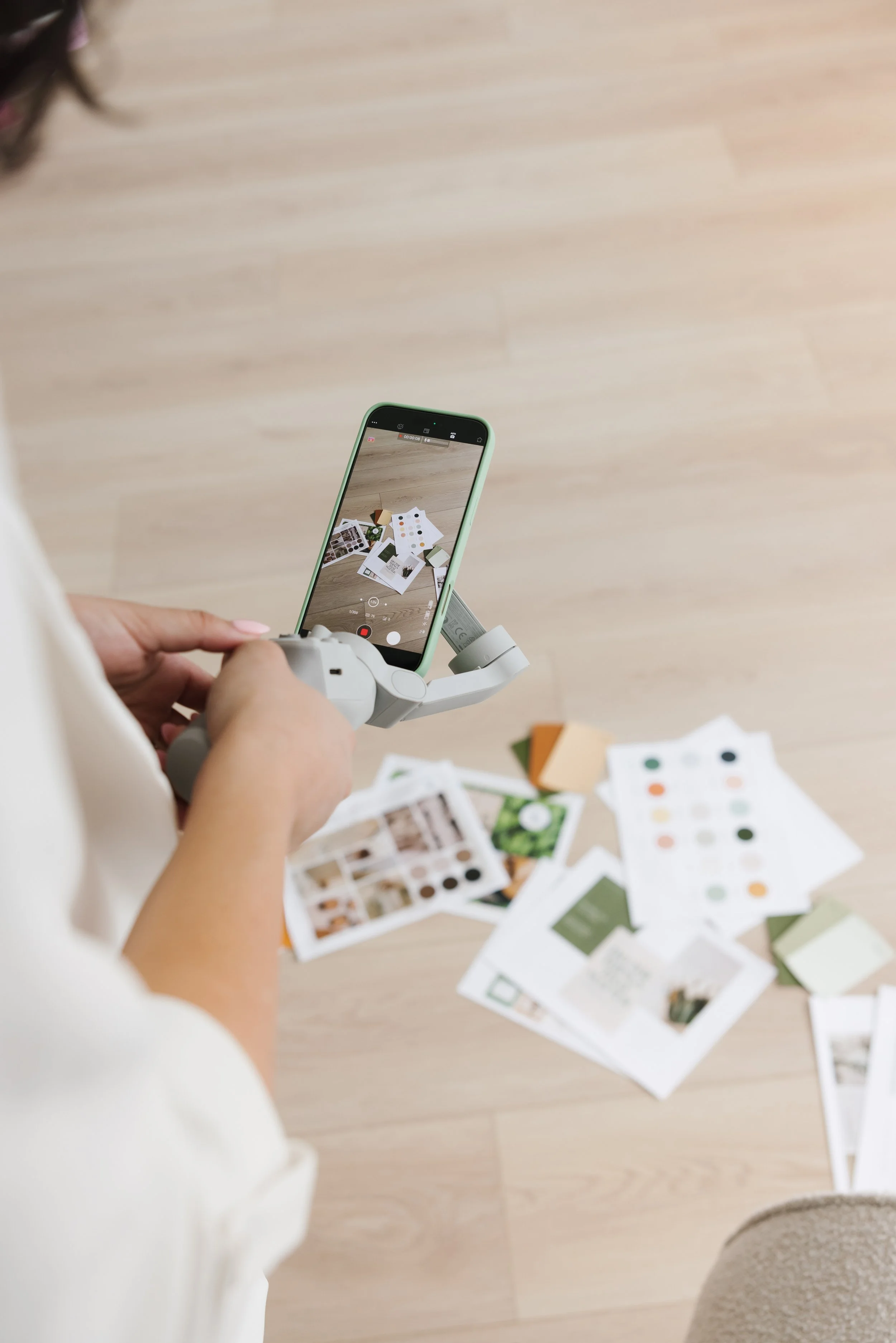 Sierra using a iphone to take a photo of colour swatches and design samples spread out on a light wooden floor.