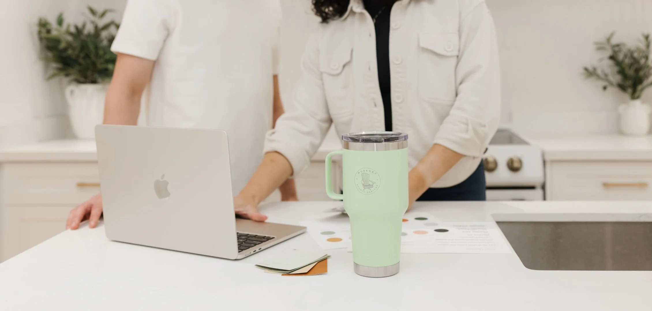 Two people stand at a kitchen counter with a laptop, color samples, and a light green tumbler.