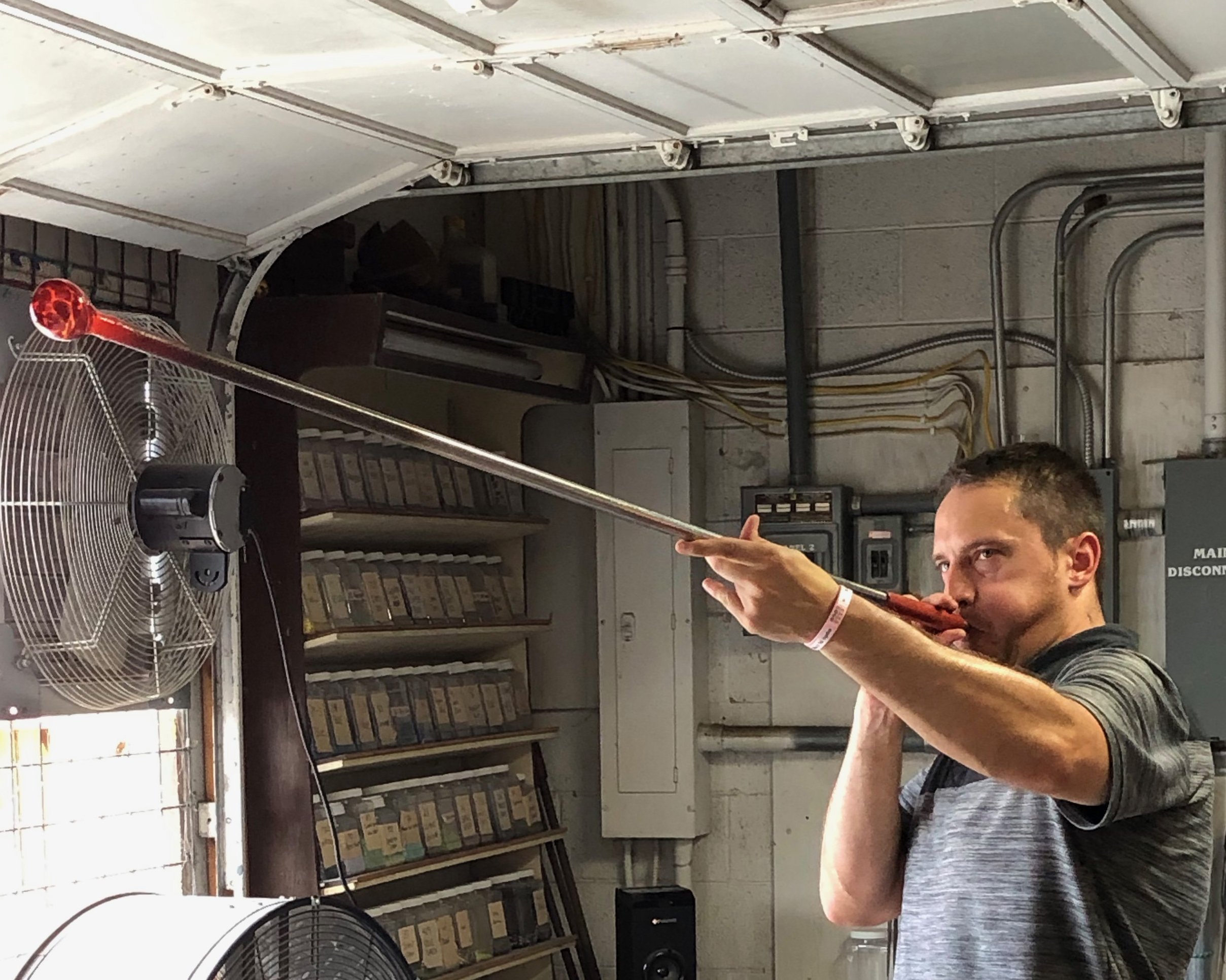 Person blowing into a glassblowing pipe with molten glass, indoors, with shelves and a fan in the background.