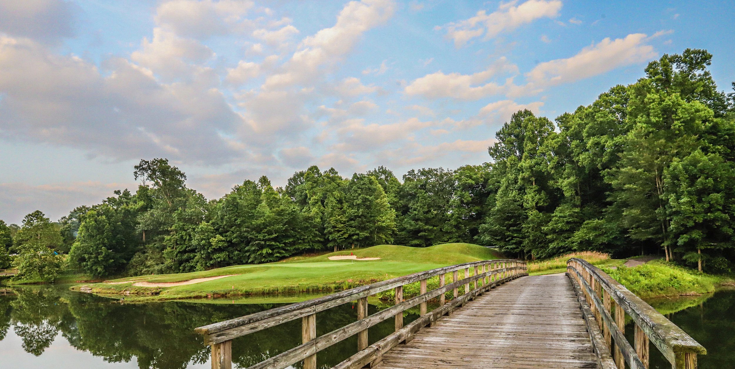 Wooden bridge over water leading to a golf course surrounded by trees, with a cloudy sky in the background.