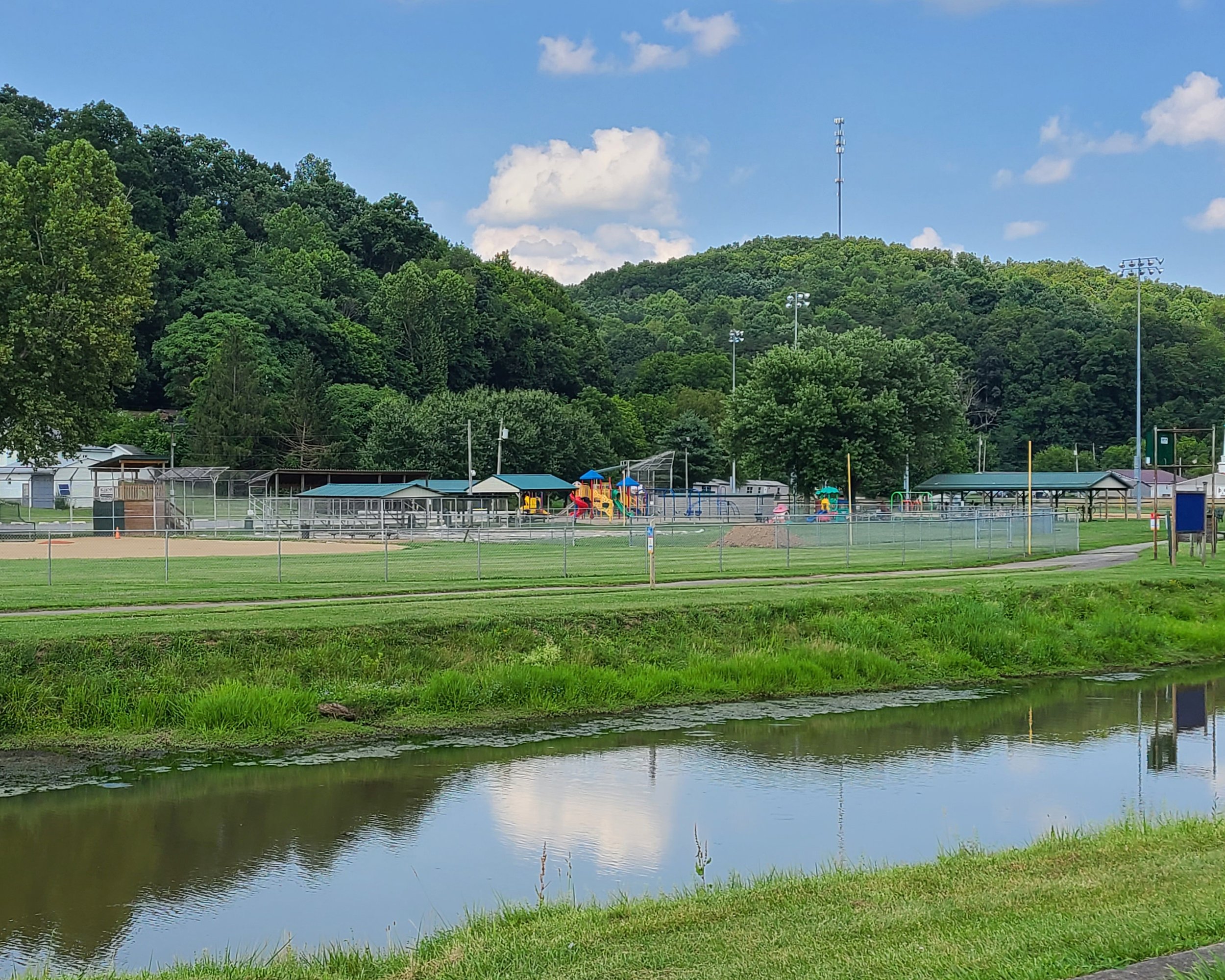 Scenic view of a park with a playground and baseball fields, surrounded by trees and hills, with a small body of water in the foreground.
