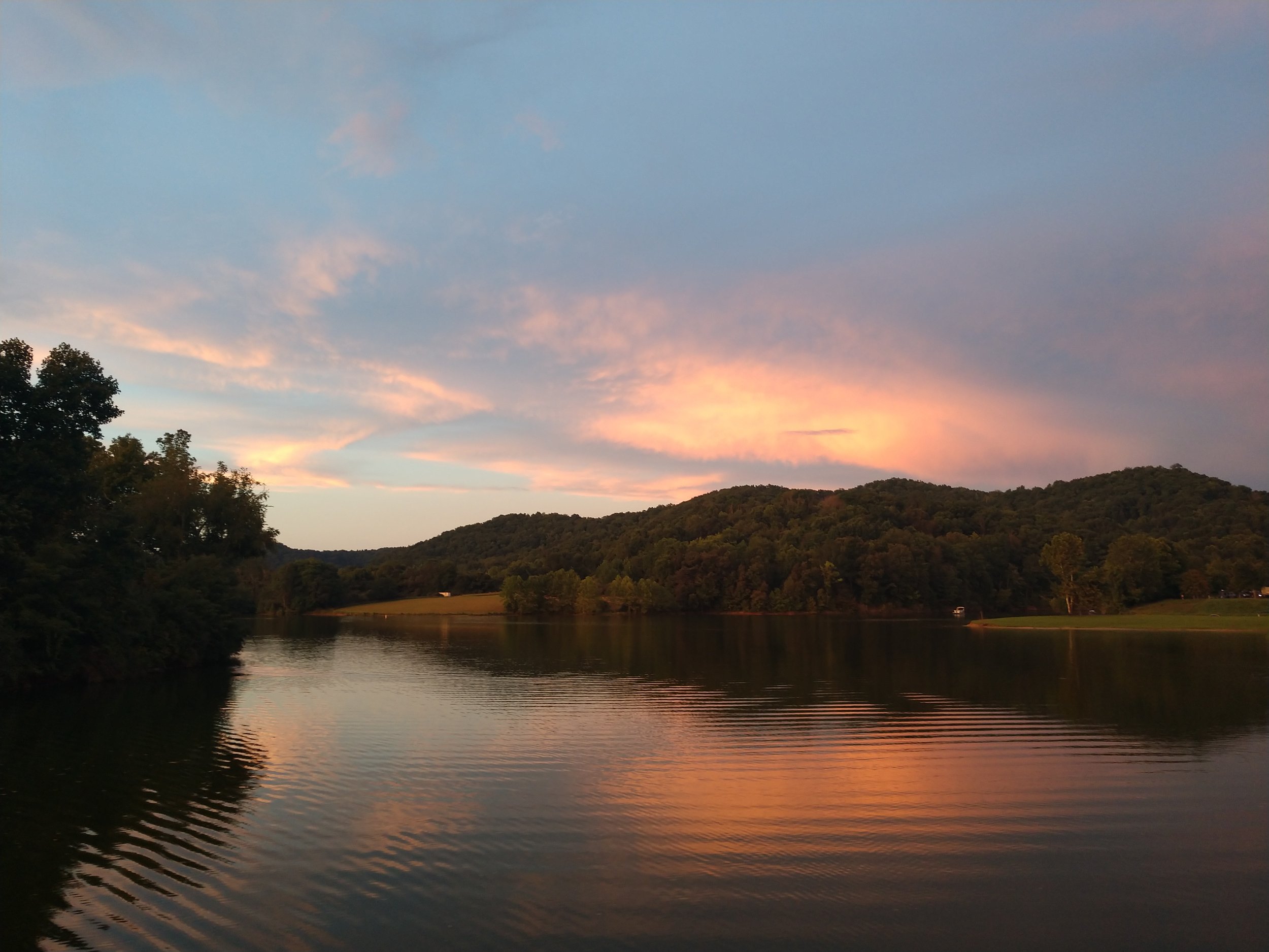 Scenic view of a lake with reflections of a colorful sunset, surrounded by trees and hills.