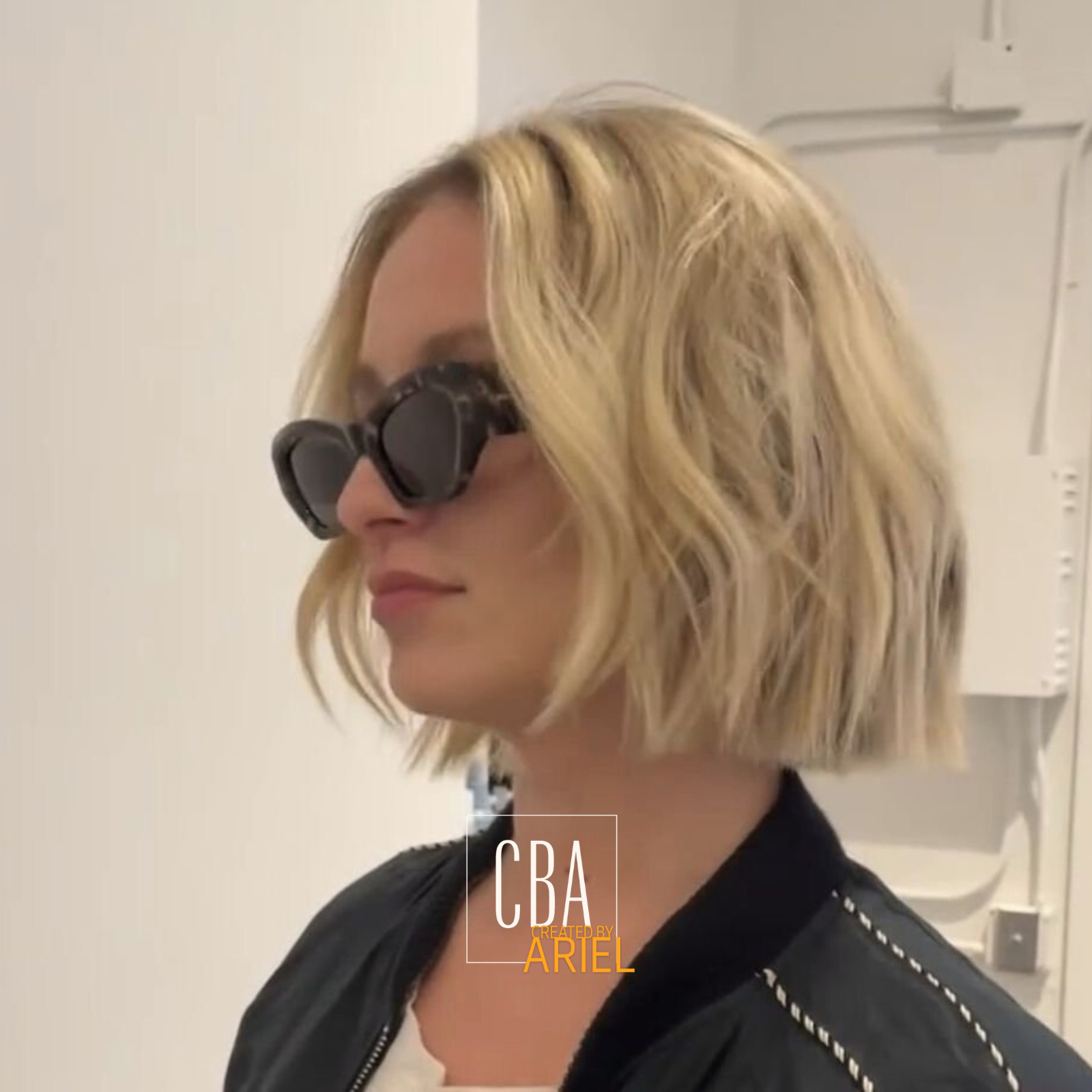 A woman with blonde, wavy, chin-length hair wearing dark sunglasses and a black jacket, standing indoors against a plain wall.