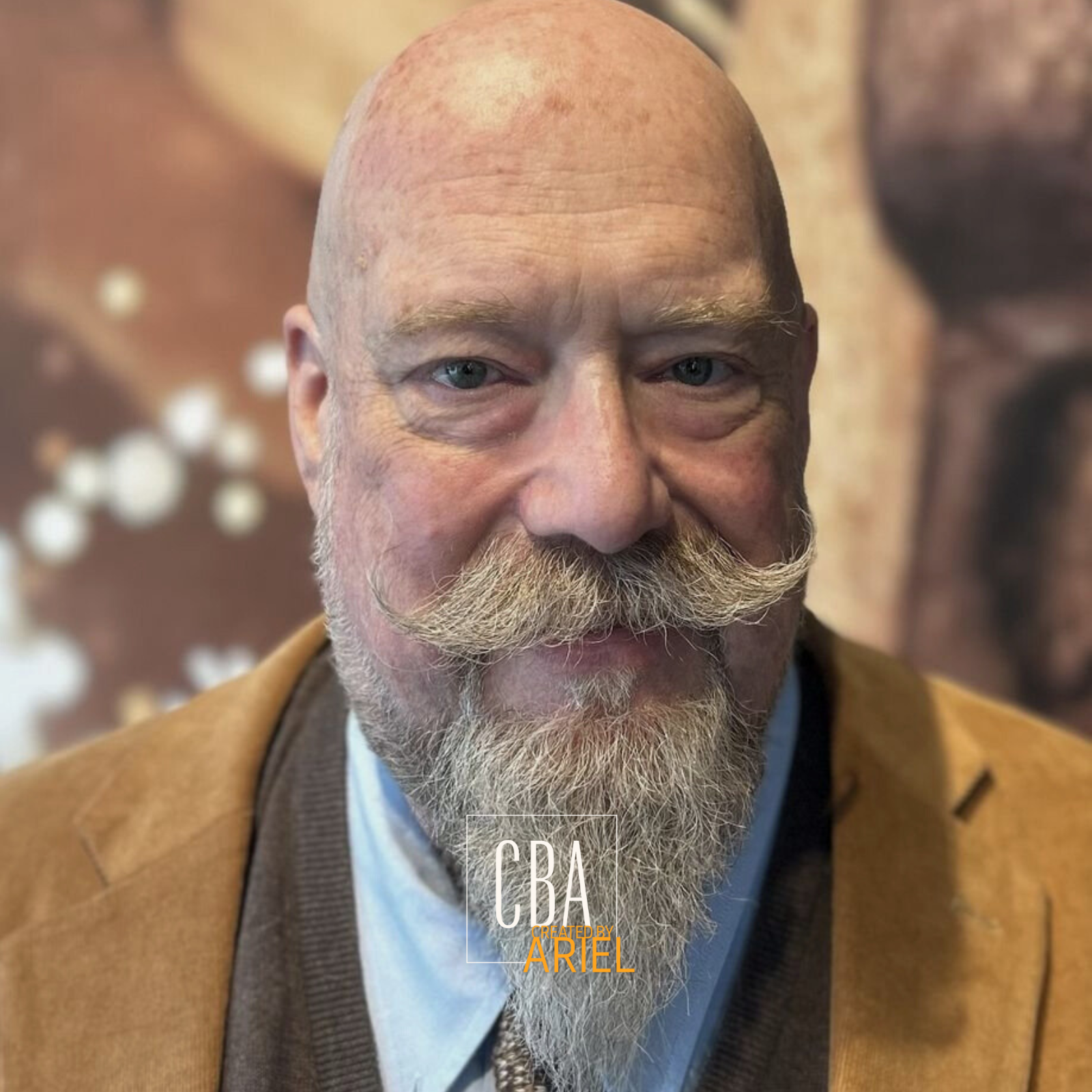 Close-up portrait of an older man with a bald head, blue eyes, a full beard, and a handlebar mustache, wearing a tan blazer over a white shirt, with a blurred background.