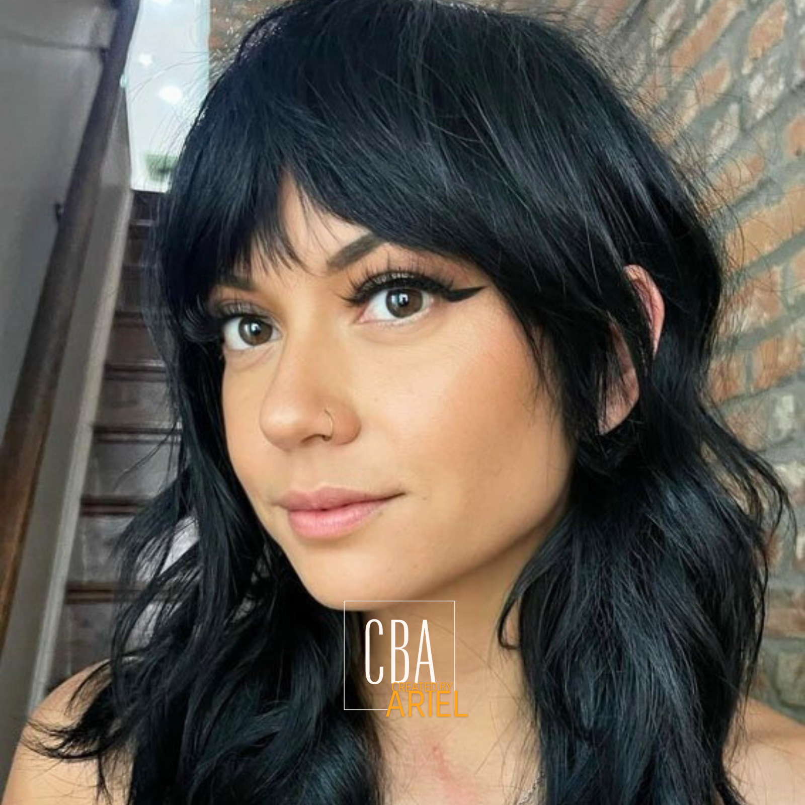 Close-up of a young woman with shoulder-length black wavy hair, wearing makeup with eyeliner, and a small nose ring, indoor with brick wall and staircase in background.