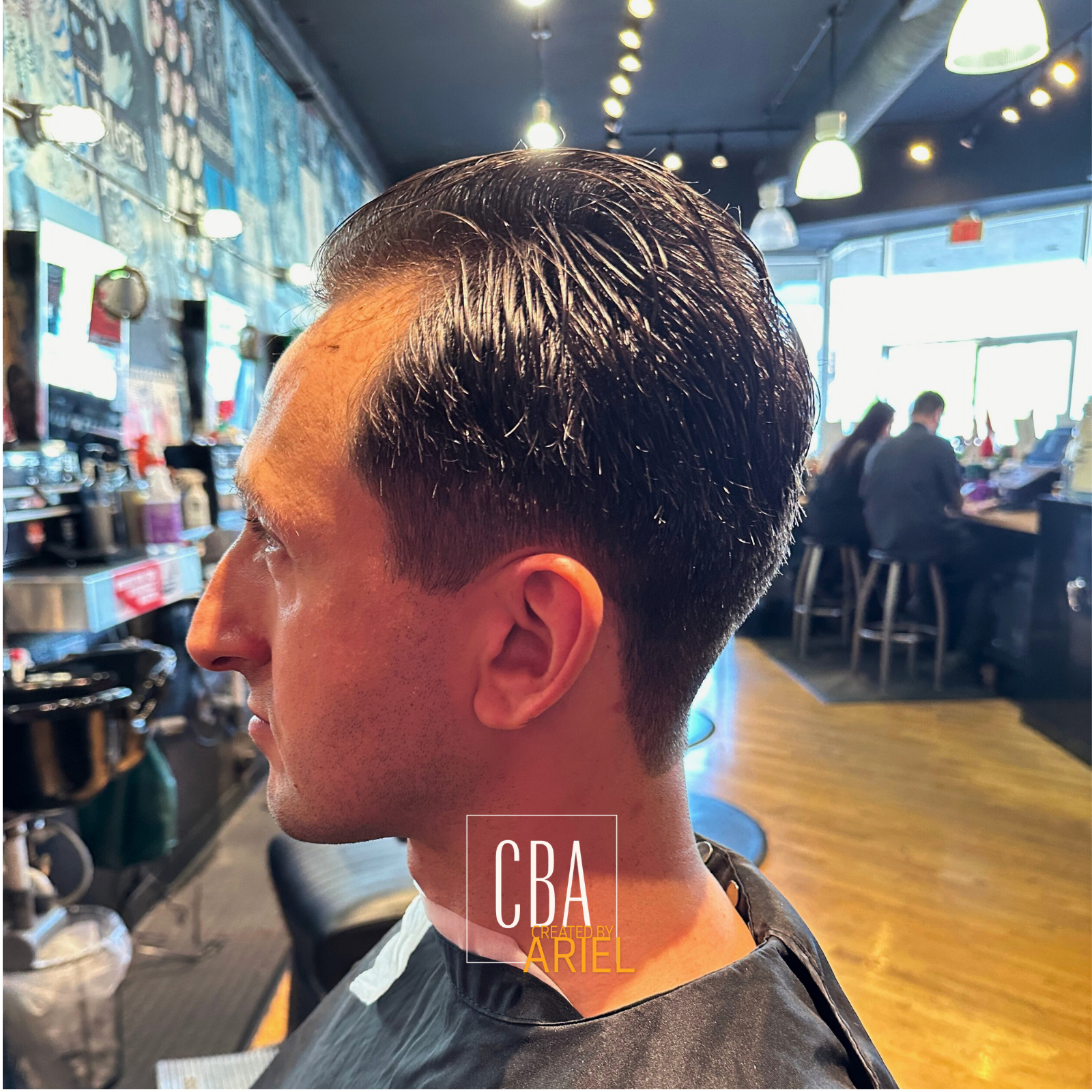 A man sitting in a barbershop with wet, slicked-back hair, wearing a black cape, with a busy cafe or restaurant background and two people sitting at a counter.