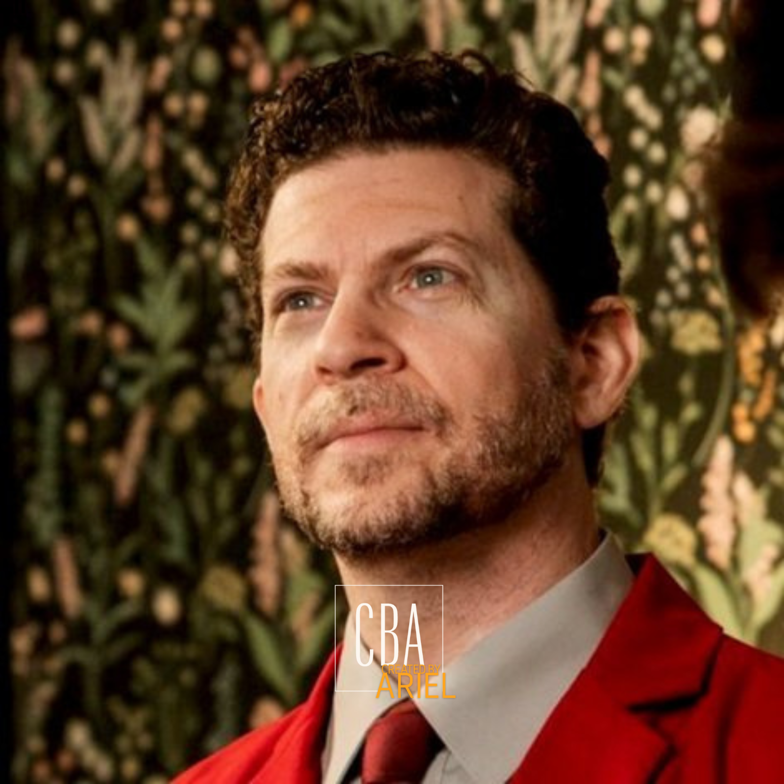 Close-up of a man with curly brown hair and a beard, wearing a red suit jacket and a gray shirt, standing in front of a leafy green background.