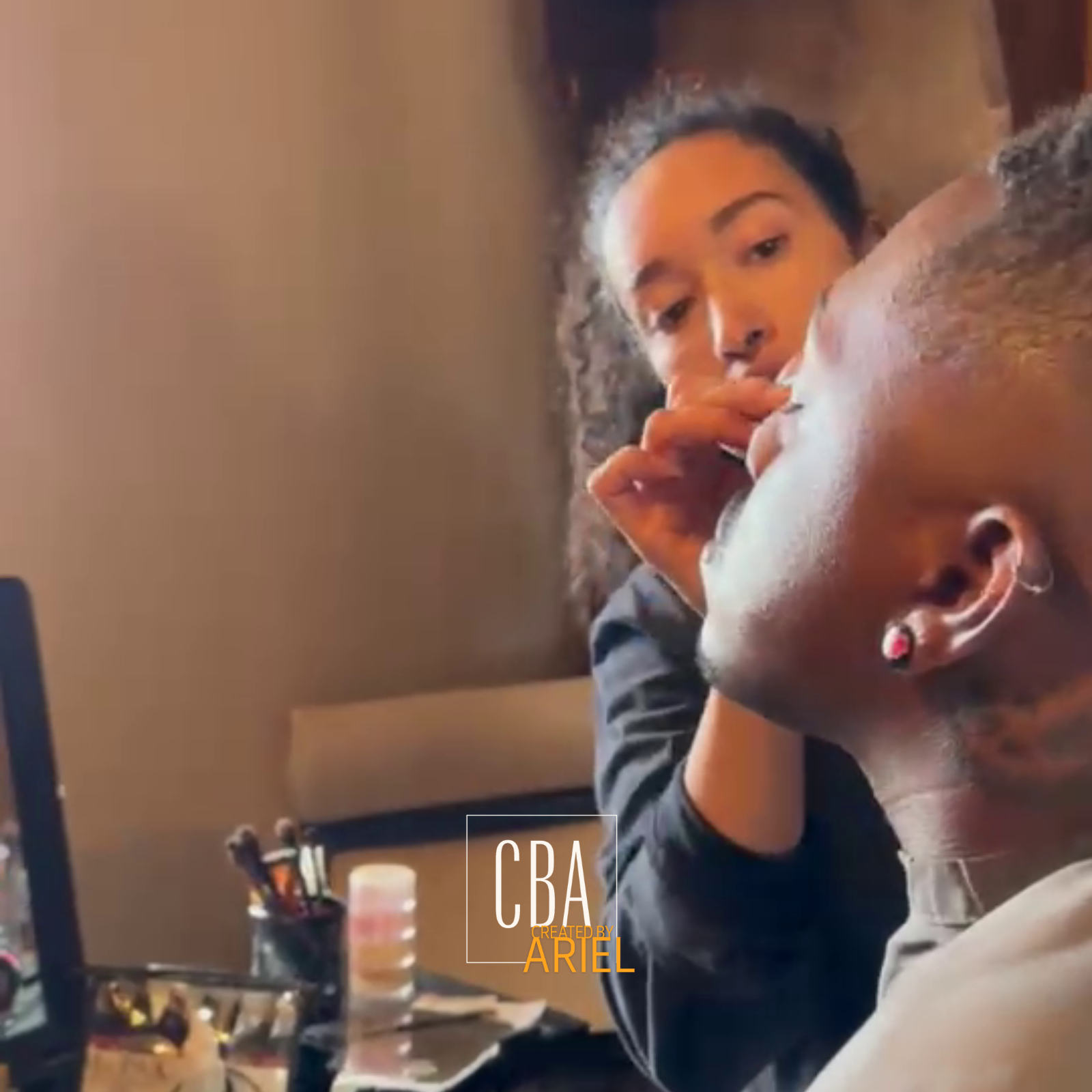 A woman applying makeup or eye makeup to a person with a shaved or bald head, sitting at a table with makeup supplies and brushes.