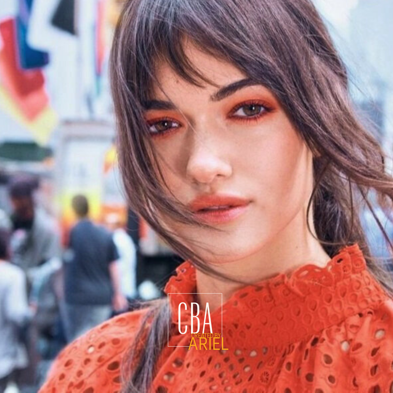 A young woman with shoulder-length brown hair and light makeup, wearing an orange eyelet blouse, posing outdoors with a blurred crowd and cityscape in the background.
